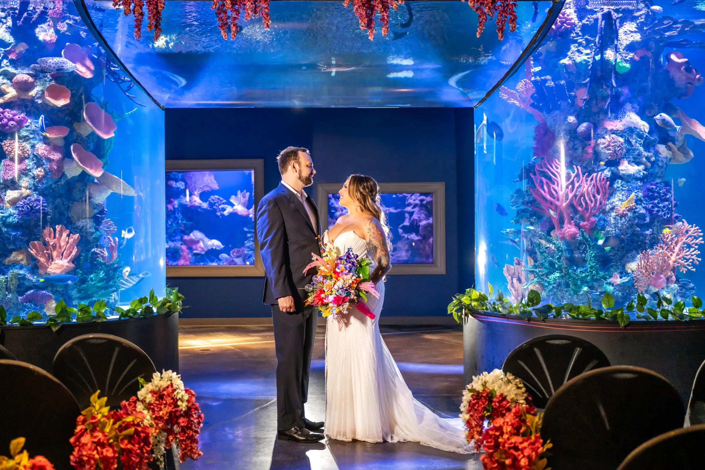 Bride and groom sharing an intimate moment during a styled wedding ceremony surrounded by vibrant coral reef aquarium tanks at Daytona Aquarium in Daytona Beach Florida, with colorful tropical bouquet and floral aisle arrangements in an alternative aquarium wedding venue setting
