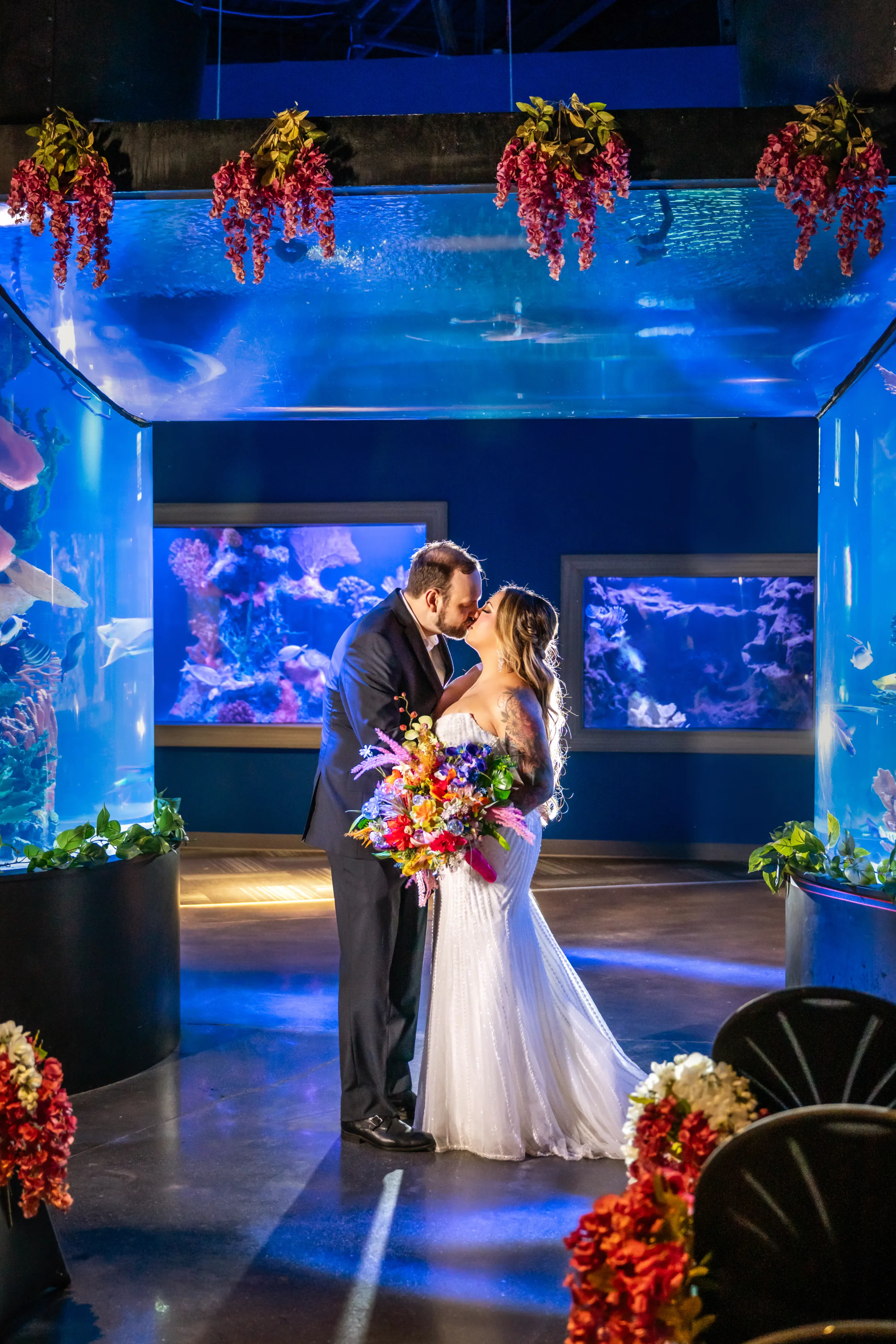 Bride and groom sharing their first kiss as a married couple surrounded by glowing blue aquarium tanks filled with tropical fish and coral at Daytona Aquarium, an alternative and colorful wedding venue in Daytona Beach Florida, with a vibrant wildflower bridal bouquet and hanging floral decor overhead