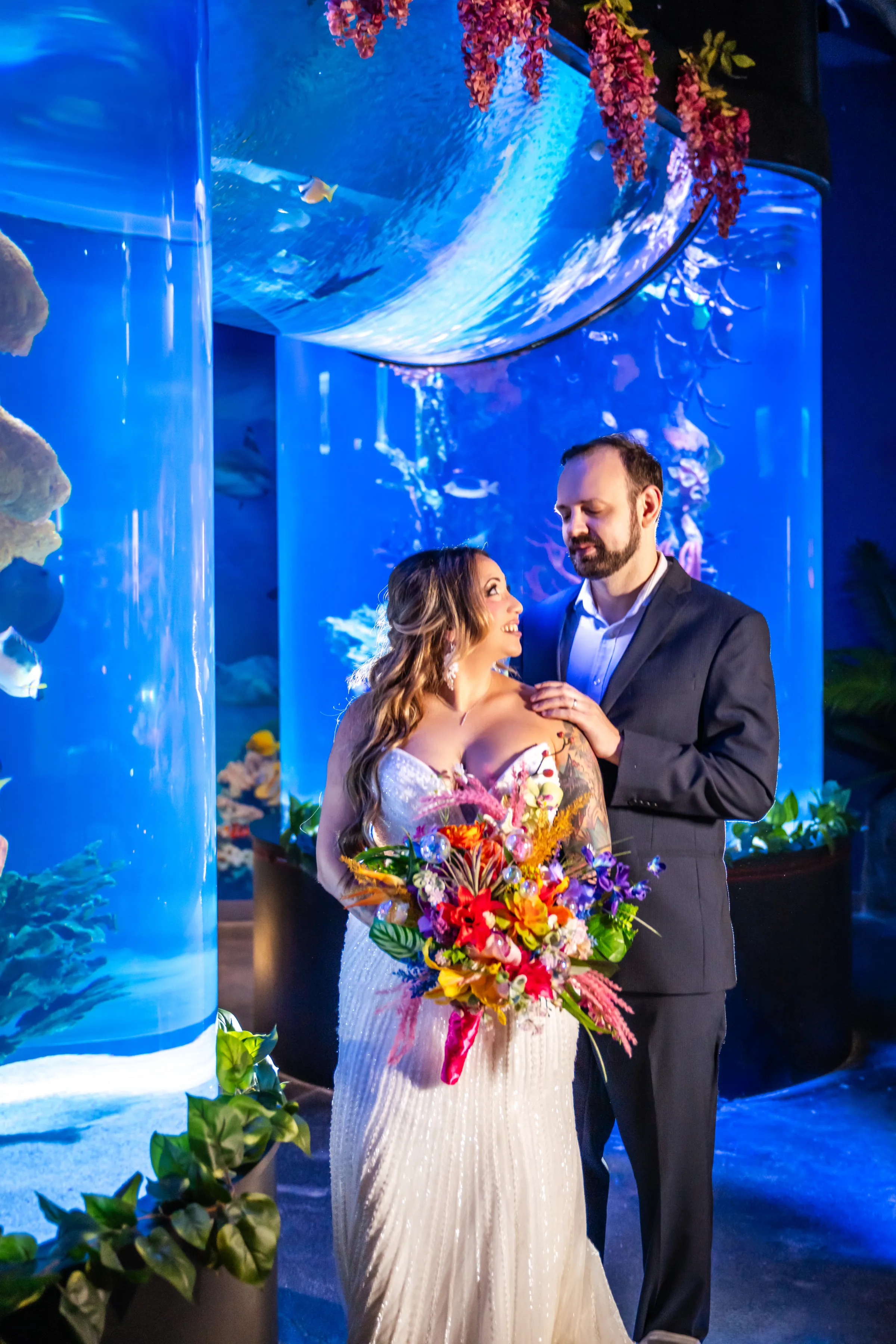 Bride and groom posing beside a vibrant coral reef aquarium with a colorful tropical wedding bouquet