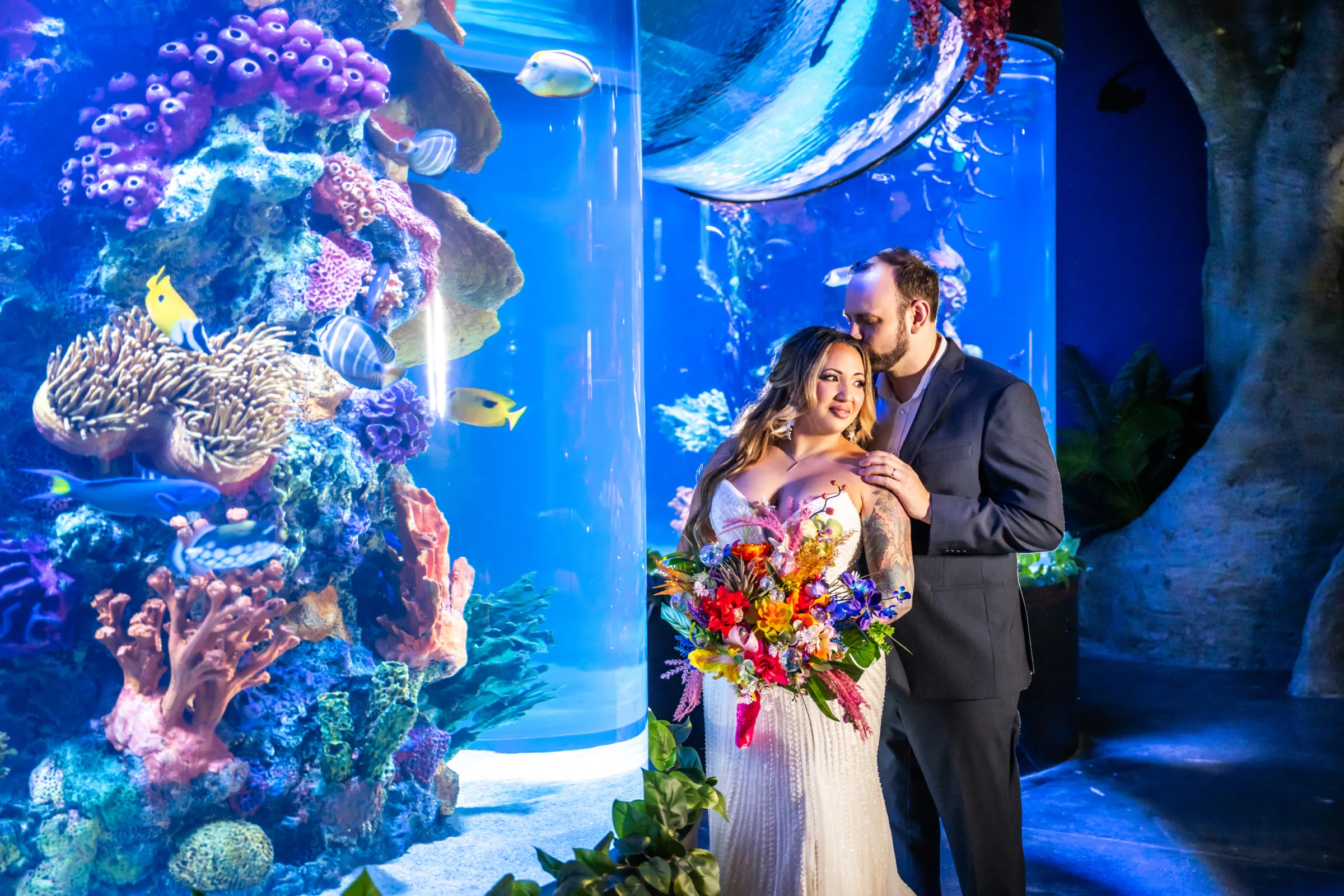 Bride and groom share a romantic moment in front of a vibrant coral reef aquarium tank at Daytona Aquarium in Daytona Beach Florida, the bride holding a colorful tropical bouquet with bold reds oranges and purples