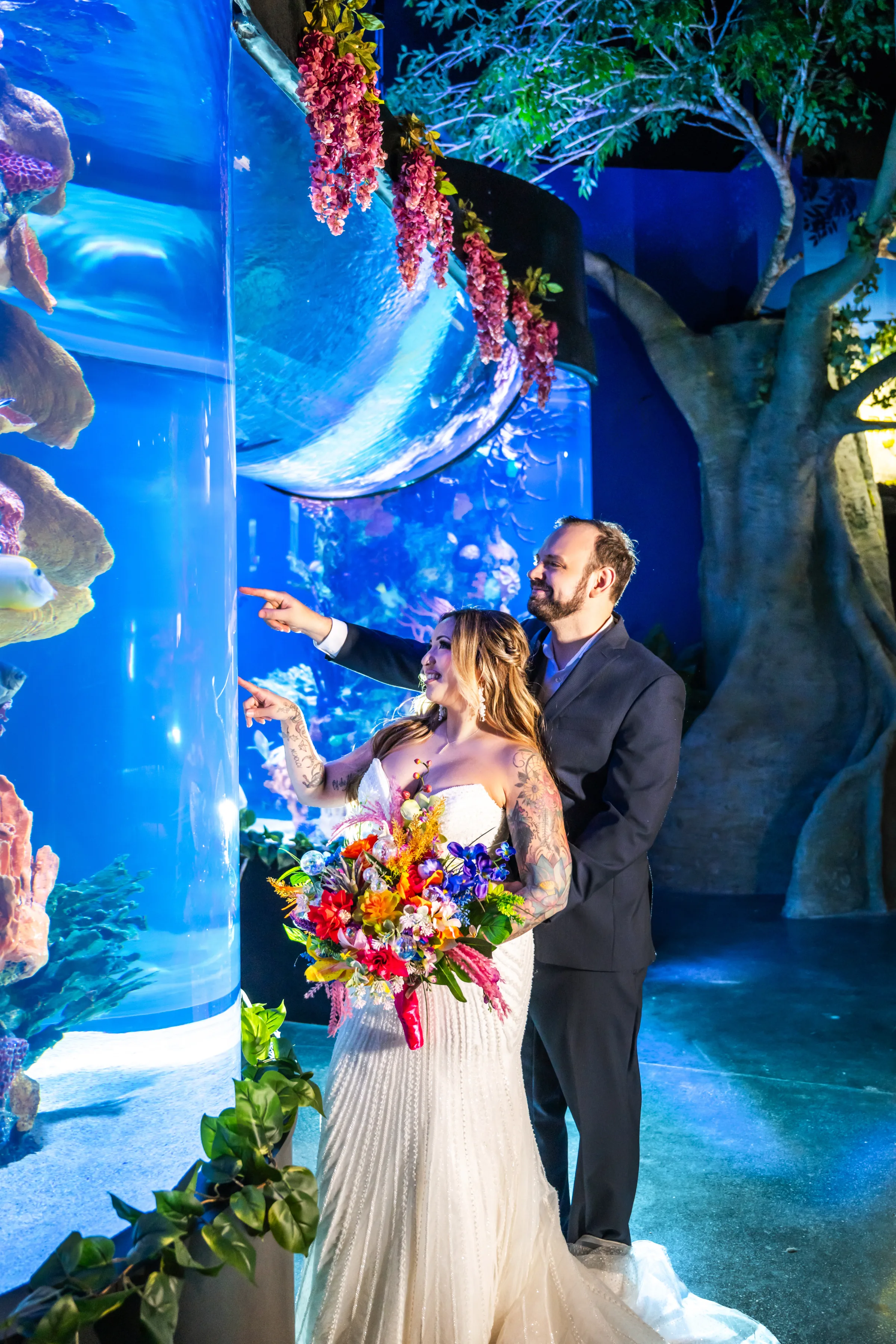 Bride and groom pointing at marine life in a cylindrical aquarium tank during a styled wedding shoot at Daytona Aquarium in Daytona Beach Florida, bride holding a vibrant tropical bouquet of red orange blue and purple blooms