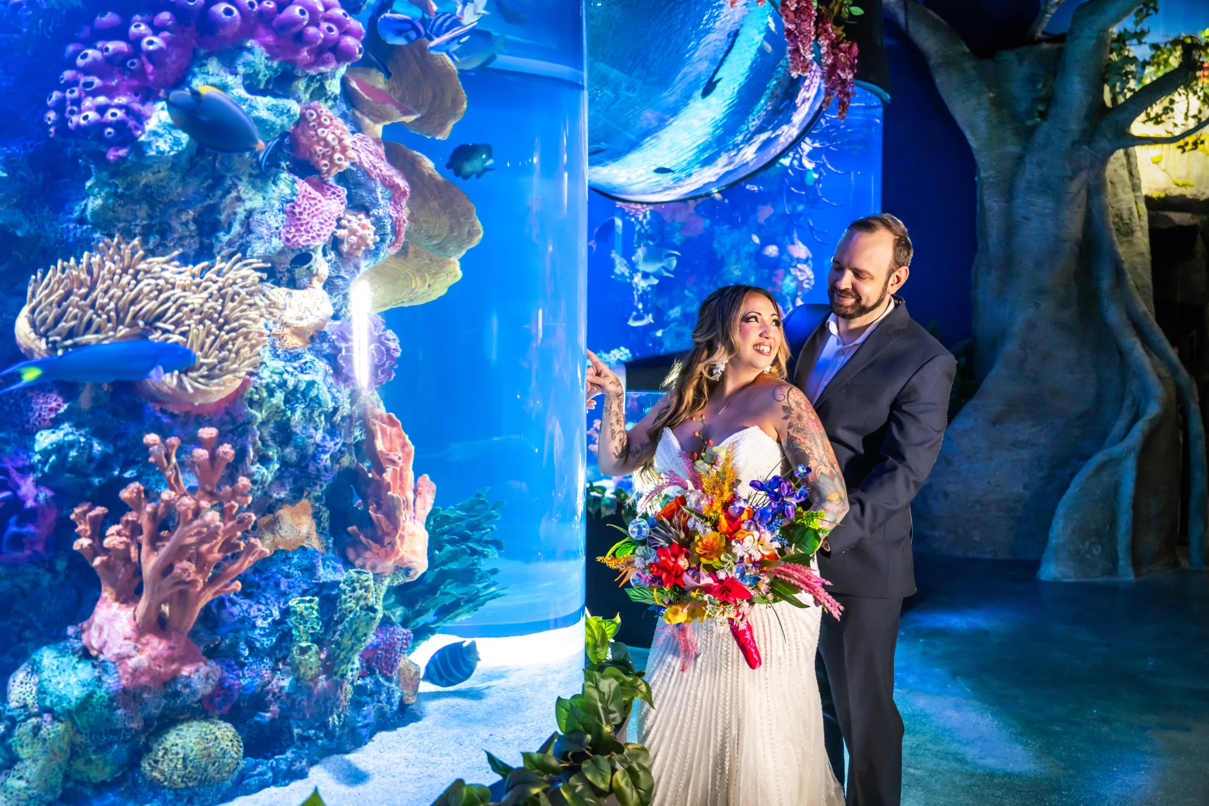 Bride with colorful tattoos and vibrant tropical bouquet pointing at coral reef display with groom during aquarium wedding styled shoot at Daytona Aquarium in Daytona Beach Florida