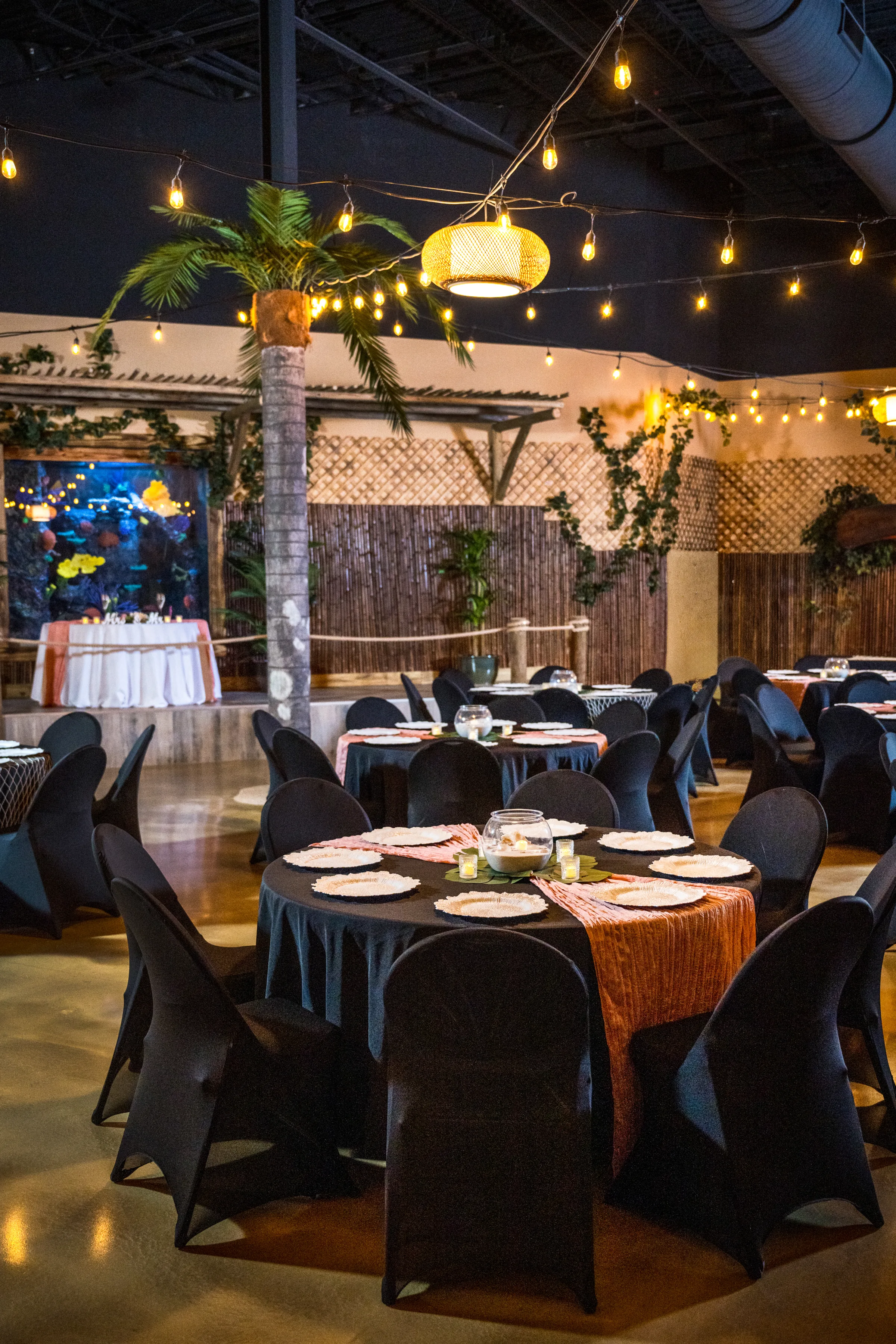 Tropical wedding reception setup at Daytona Aquarium featuring black spandex chairs, dark linen round tables with terracotta runners, white charger plates, candle centerpieces, string lights, a woven pendant light, palm tree decor, bamboo wall panels, and ivy greenery in an alternative aquarium venue setting in Daytona Beach Florida