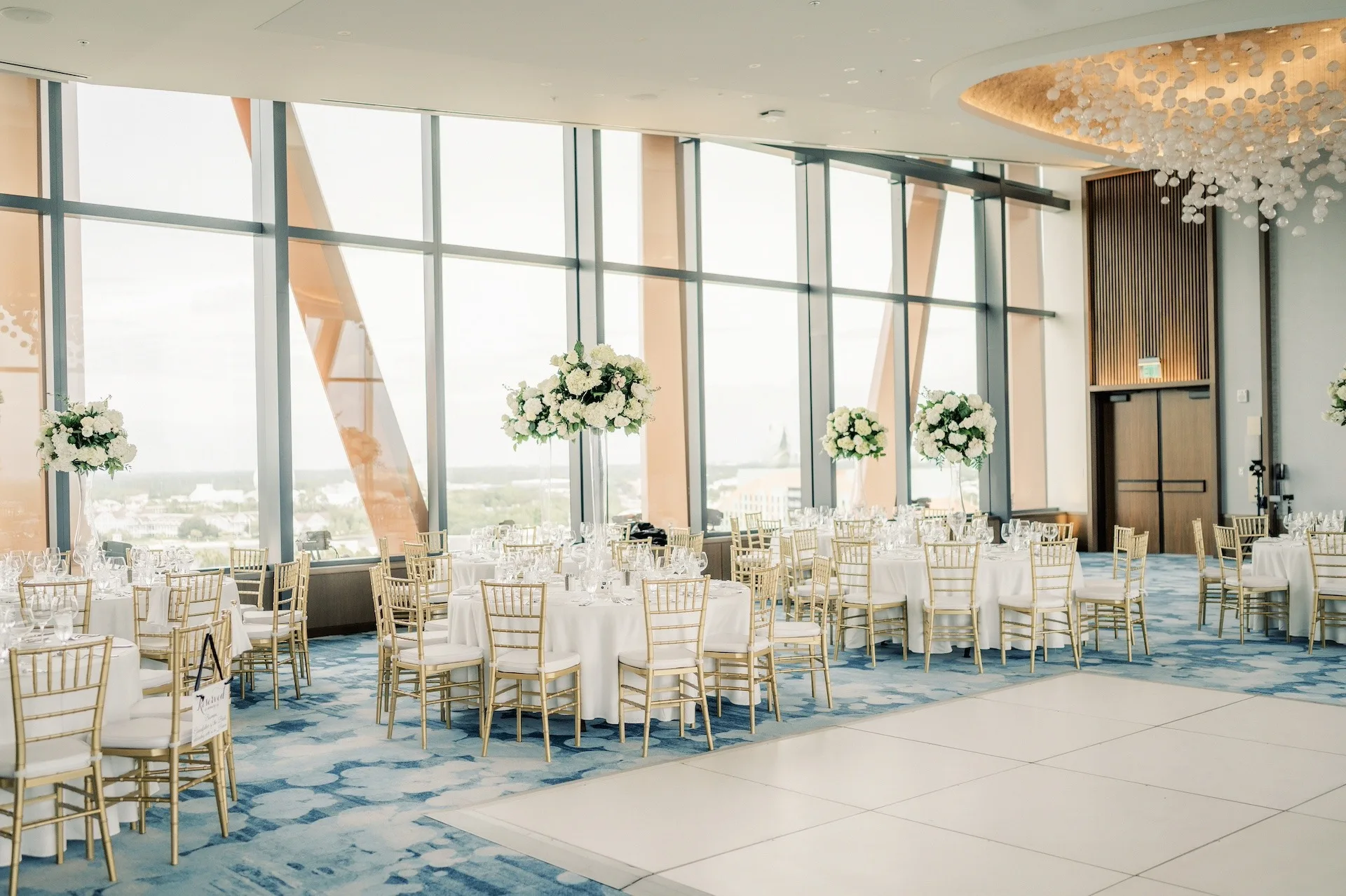 ballroom central florida wedding venue walt disney world swan reserve - Wedding Venues Collections Elegant wedding reception ballroom with floor-to-ceiling windows, gold chiavari chairs, white round tables, and tall white floral centerpieces overlooking a scenic view at Walt Disney World Swan Reserve in Central Florida