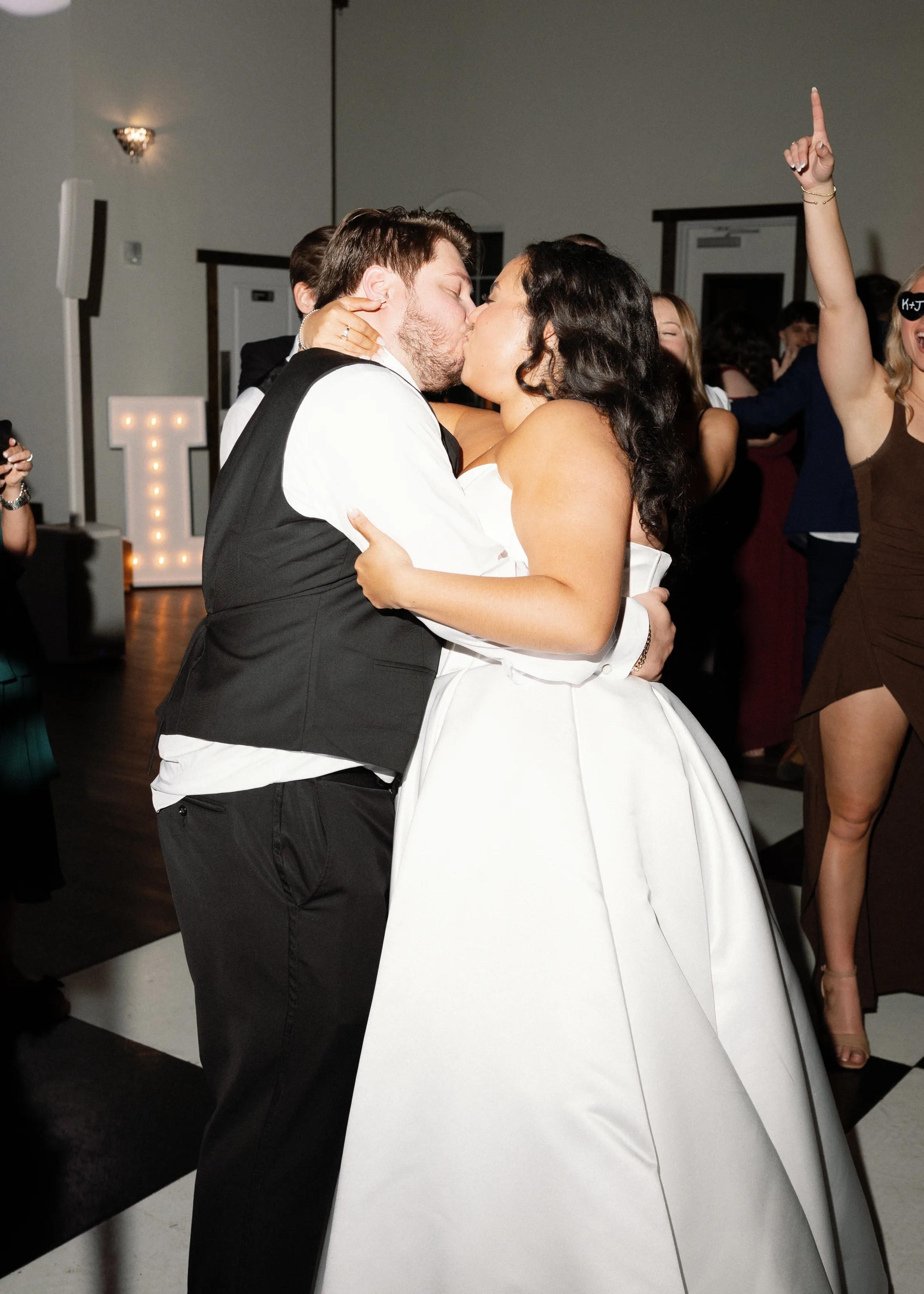 Bride in a strapless white ball gown and groom in a black tuxedo vest sharing a kiss on the dance floor surrounded by celebrating guests at a Central Florida wedding reception