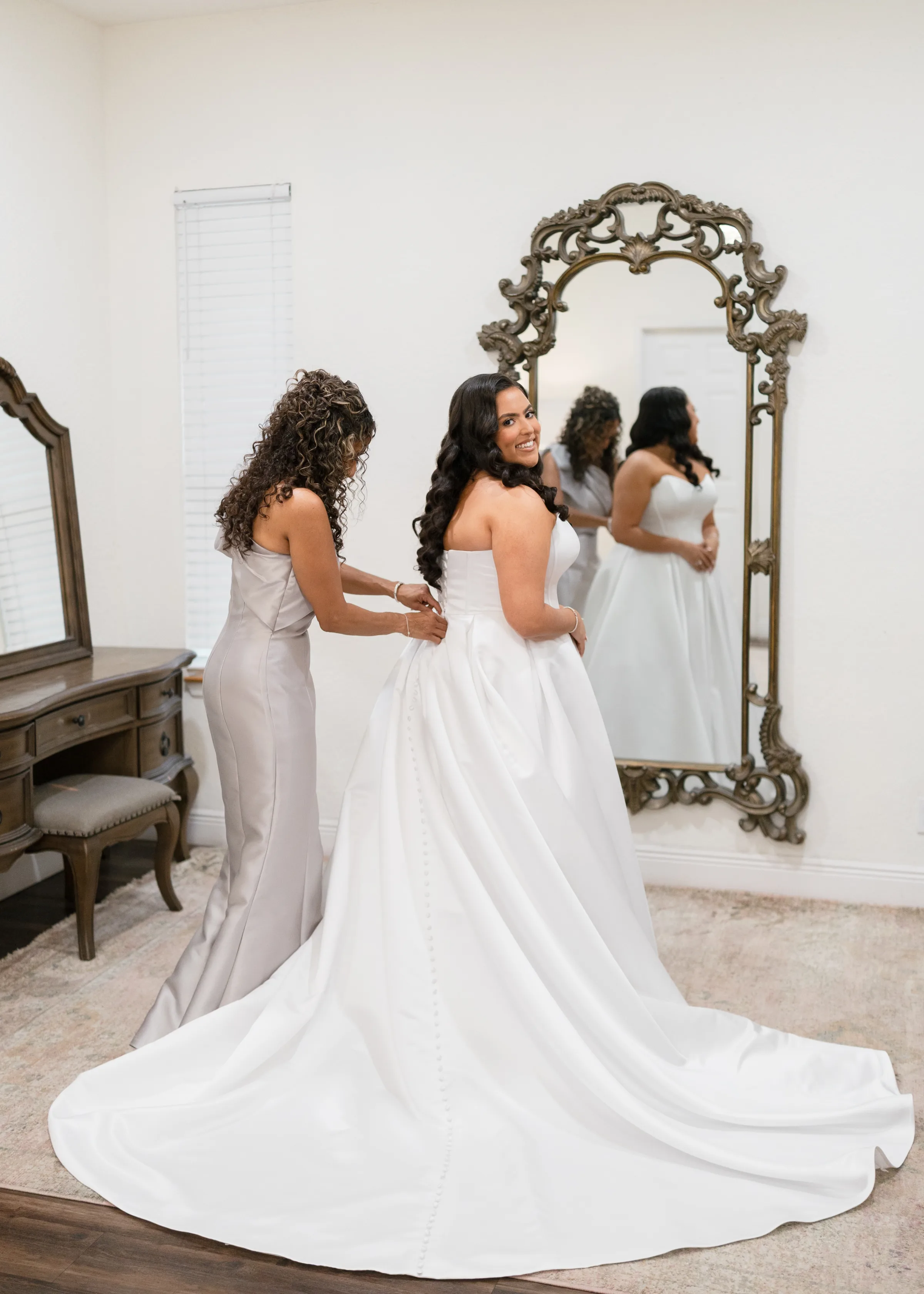 Bride smiling over her shoulder while bridesmaid fastens her strapless white ball gown in an elegant bridal suite with ornate gold mirror in Orlando, Florida