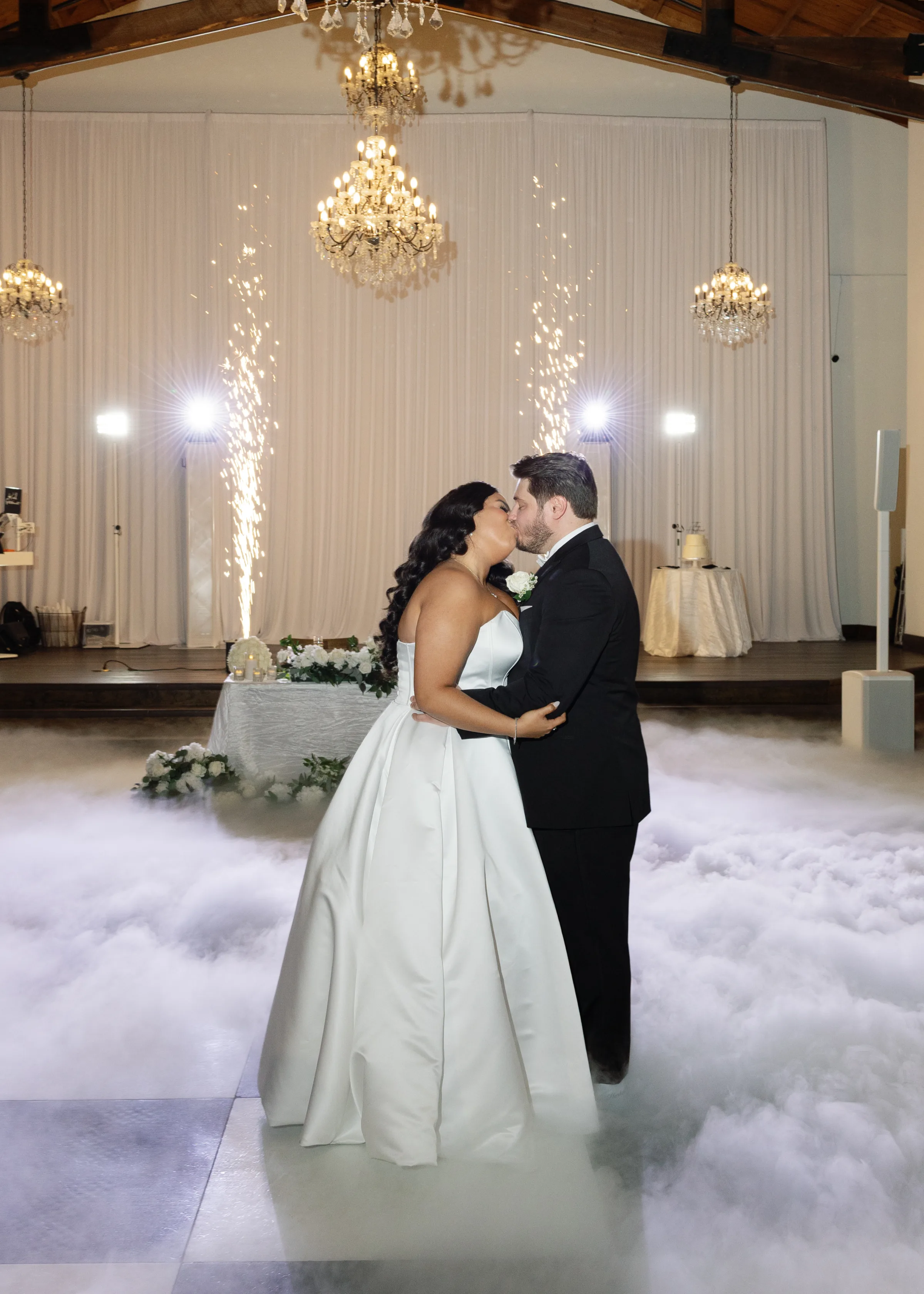 Bride and groom share a kiss during their first dance surrounded by dramatic dry ice clouds and cold sparkler fountains at an elegant Orlando wedding venue with crystal chandeliers