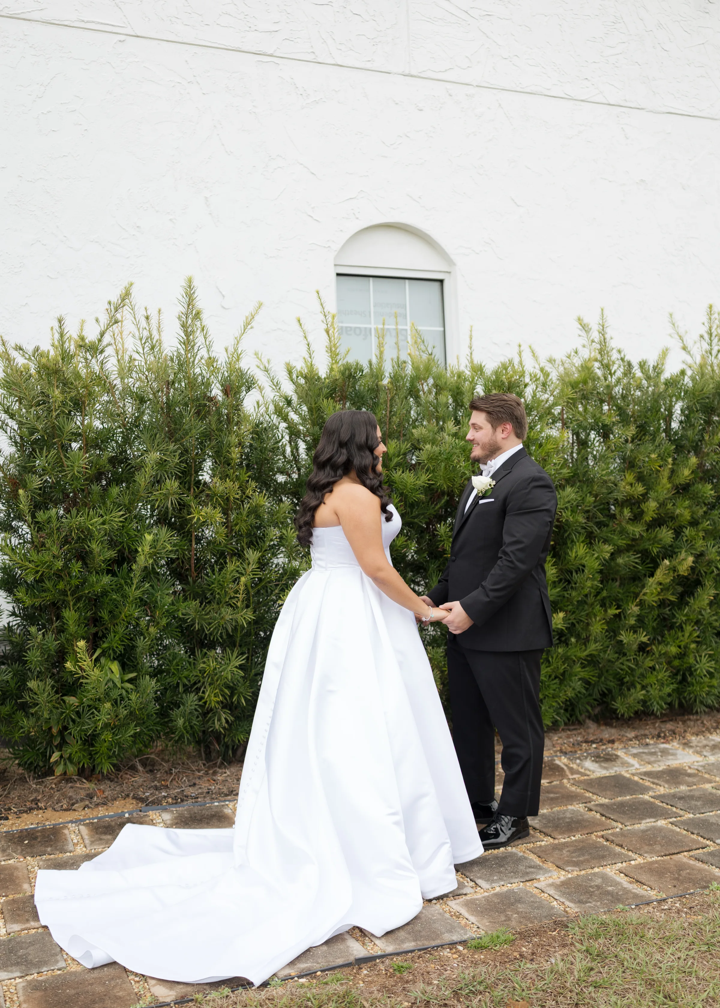 Bride in a strapless white ball gown and groom in a black tuxedo share a first look moment while holding hands outside a white church in Central Florida