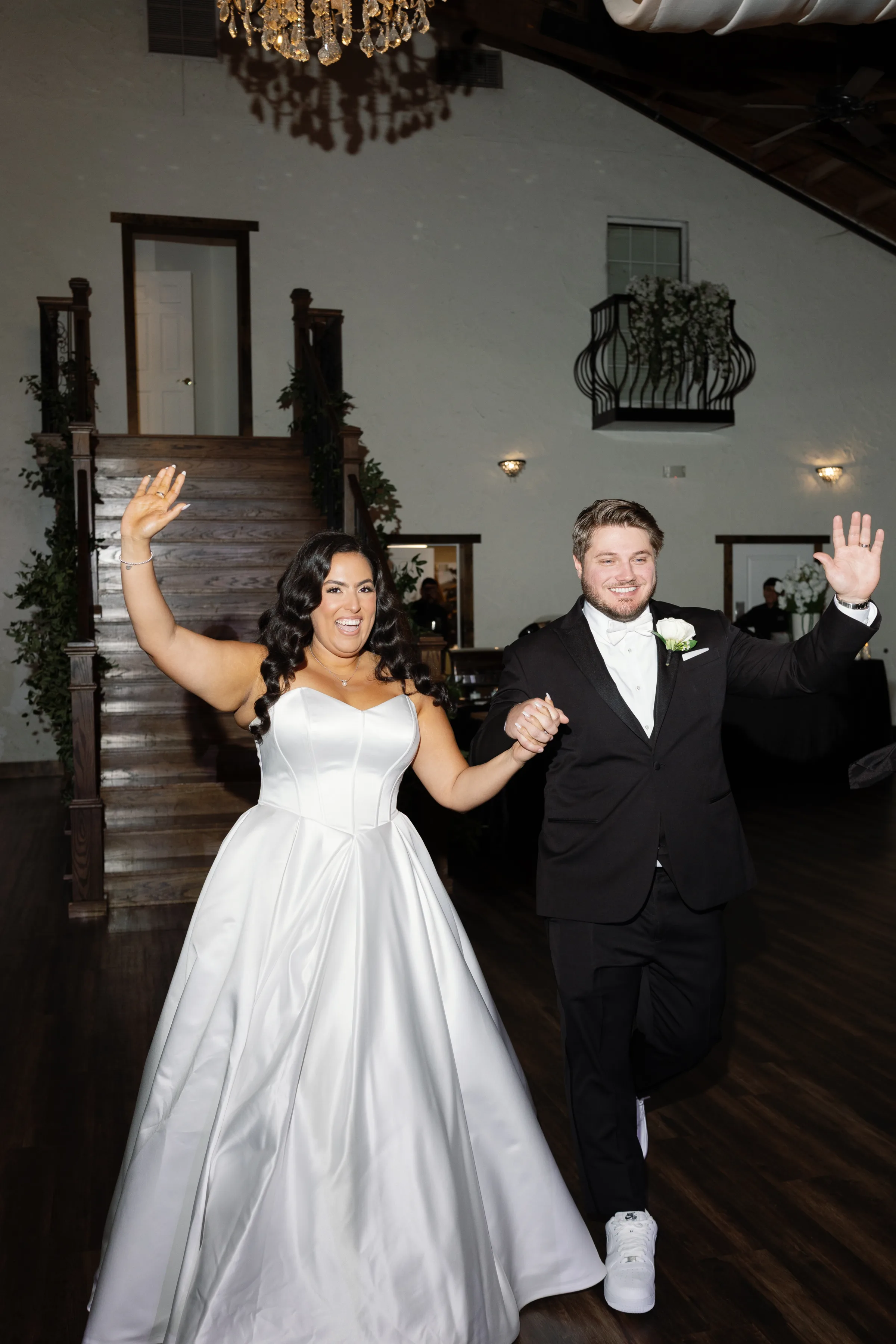 Bride in a strapless white ball gown and groom in a black tuxedo wave to guests during their grand entrance at an elegant Orlando wedding reception venue with dark wood staircase and crystal chandelier