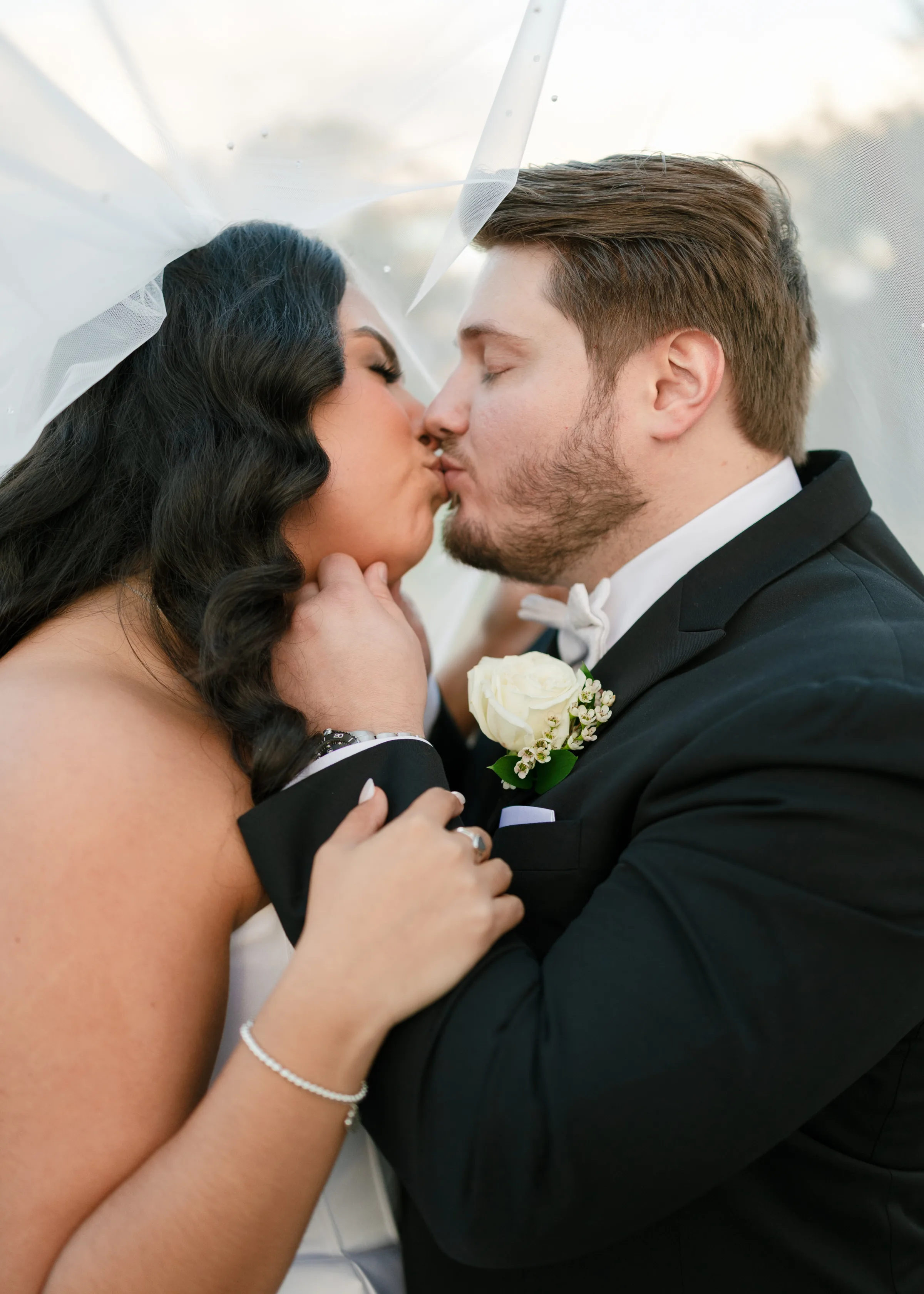 Bride and groom sharing a romantic kiss under the wedding veil at an Orlando Central Florida wedding, with the groom wearing a black tuxedo with white rose boutonniere