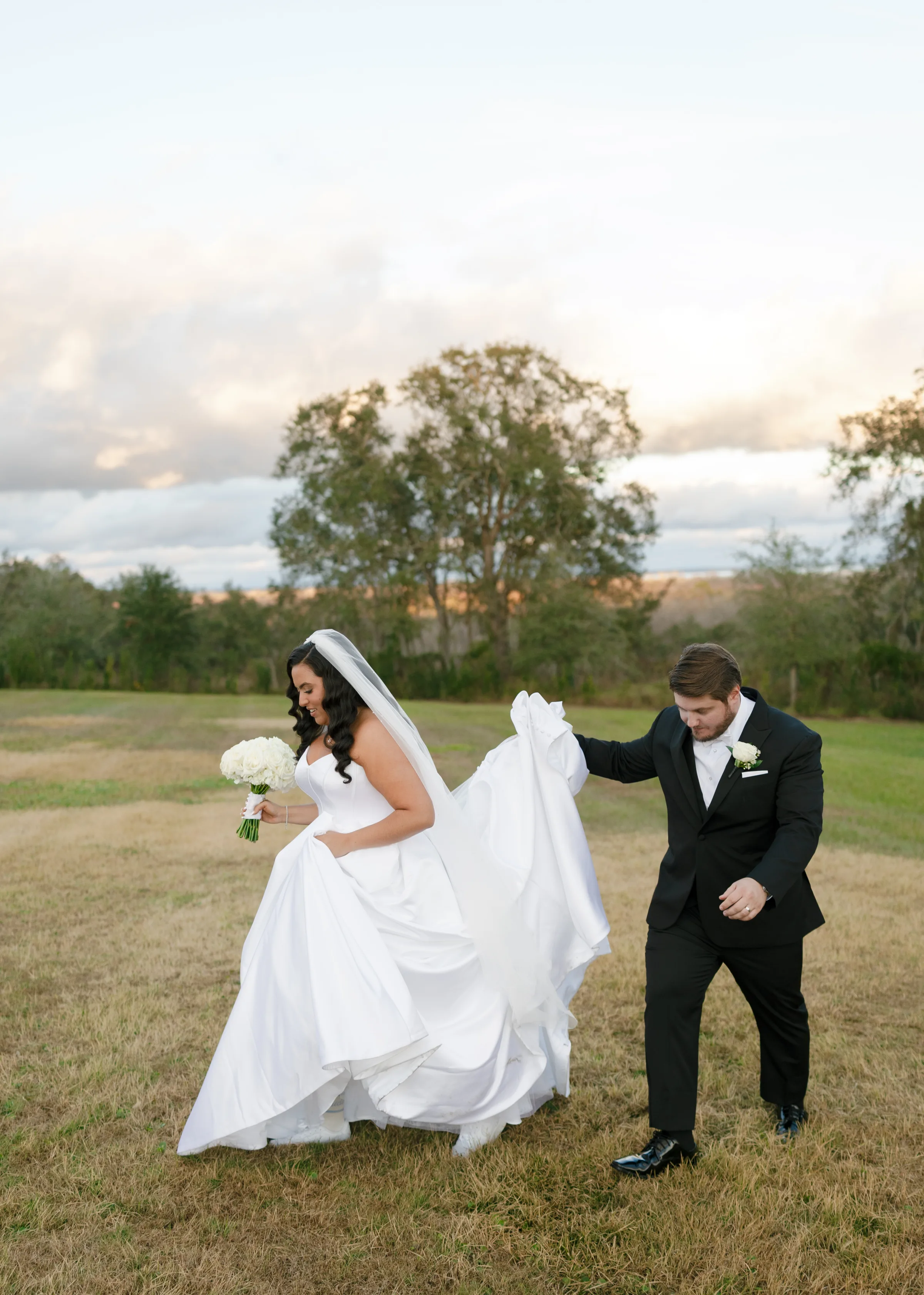 Groom in black tuxedo helping lift bride's white ball gown train during outdoor portrait session at a Central Florida wedding venue at golden hour