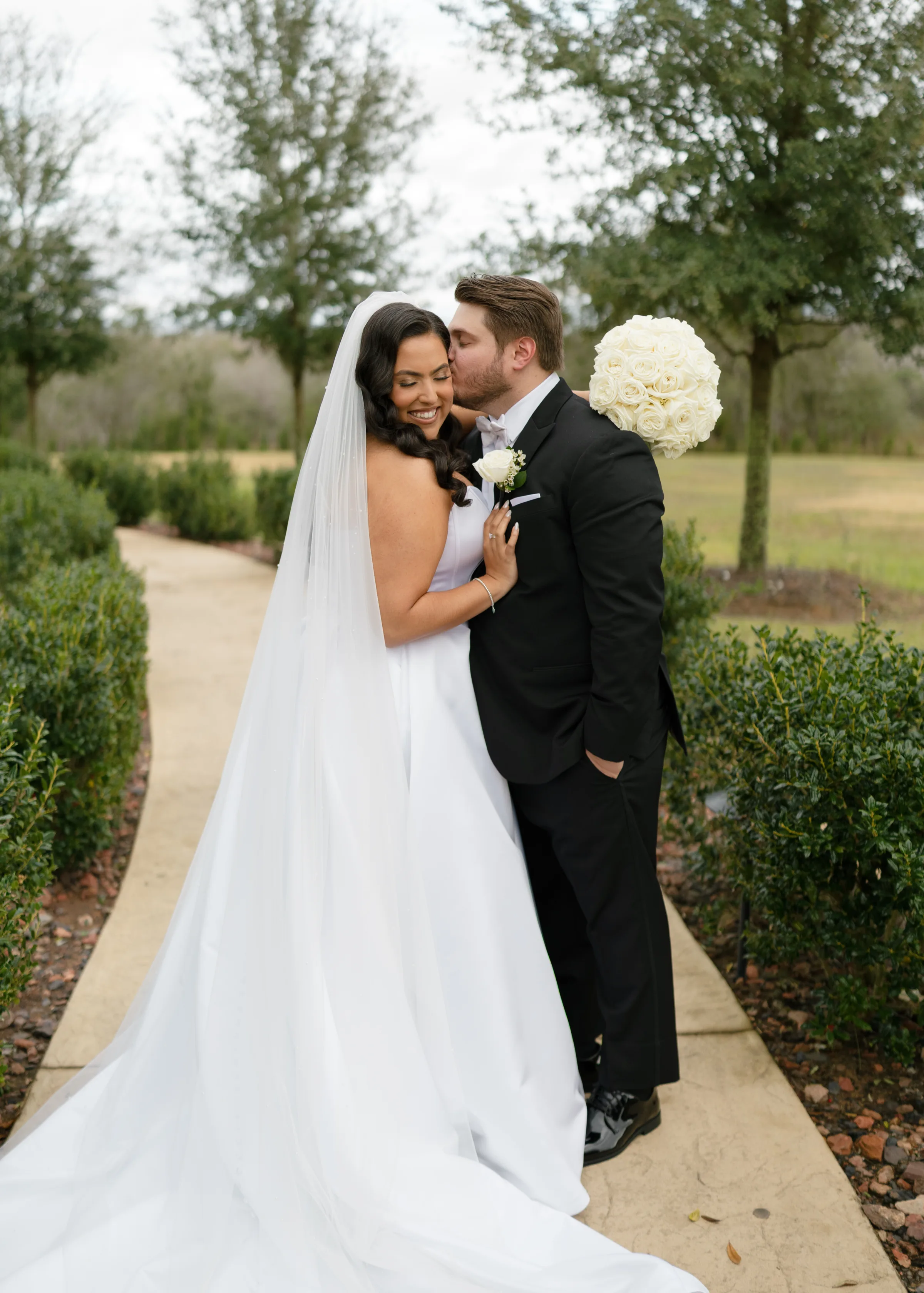 Bride in white gown with cathedral veil and white rose bouquet smiles as groom in black tuxedo kisses her cheek on a garden pathway at a Central Florida wedding venue