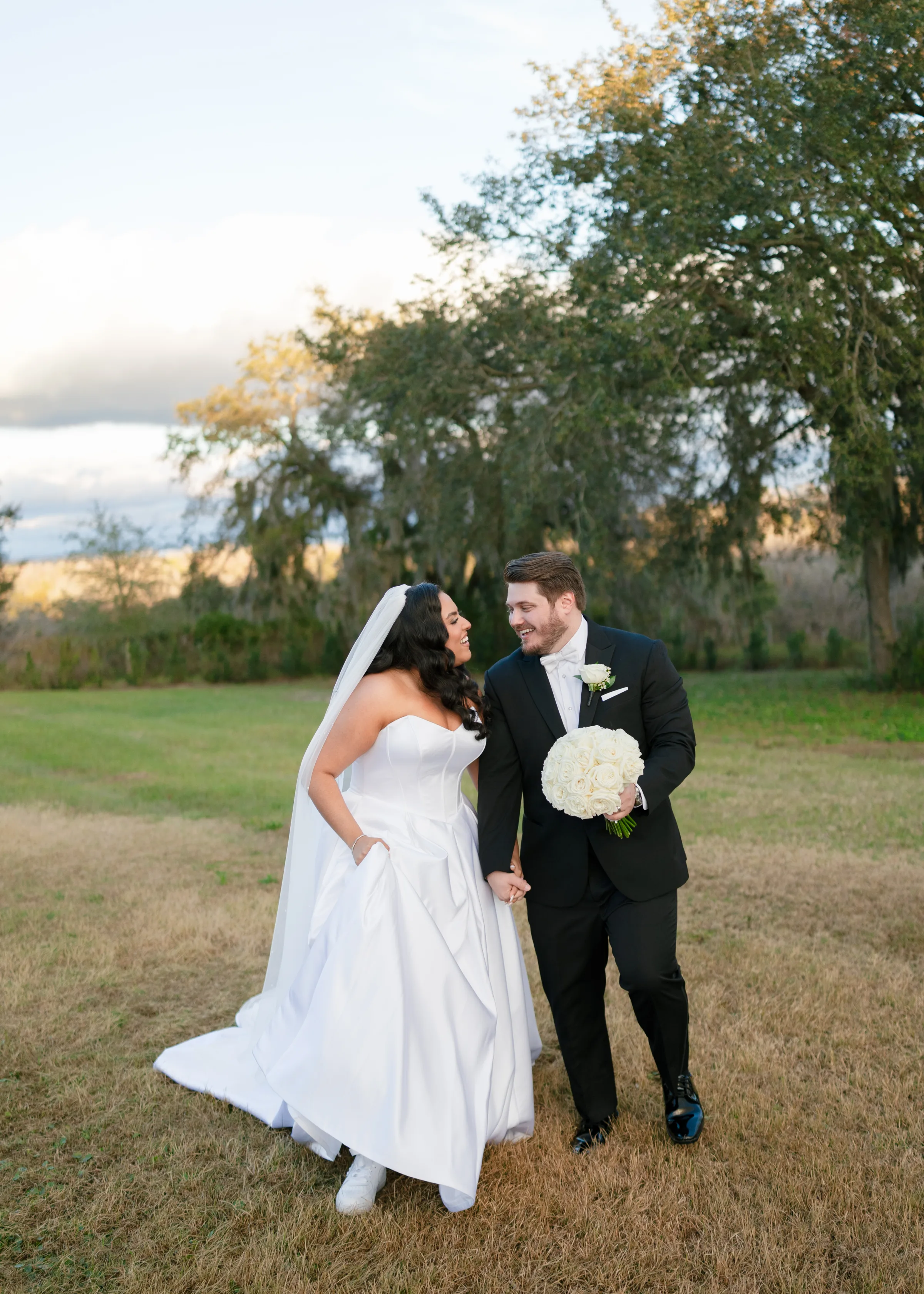 Bride in strapless white ballgown with veil and groom in black tuxedo holding white rose bouquet walk together laughing in an open field at a Central Florida wedding venue