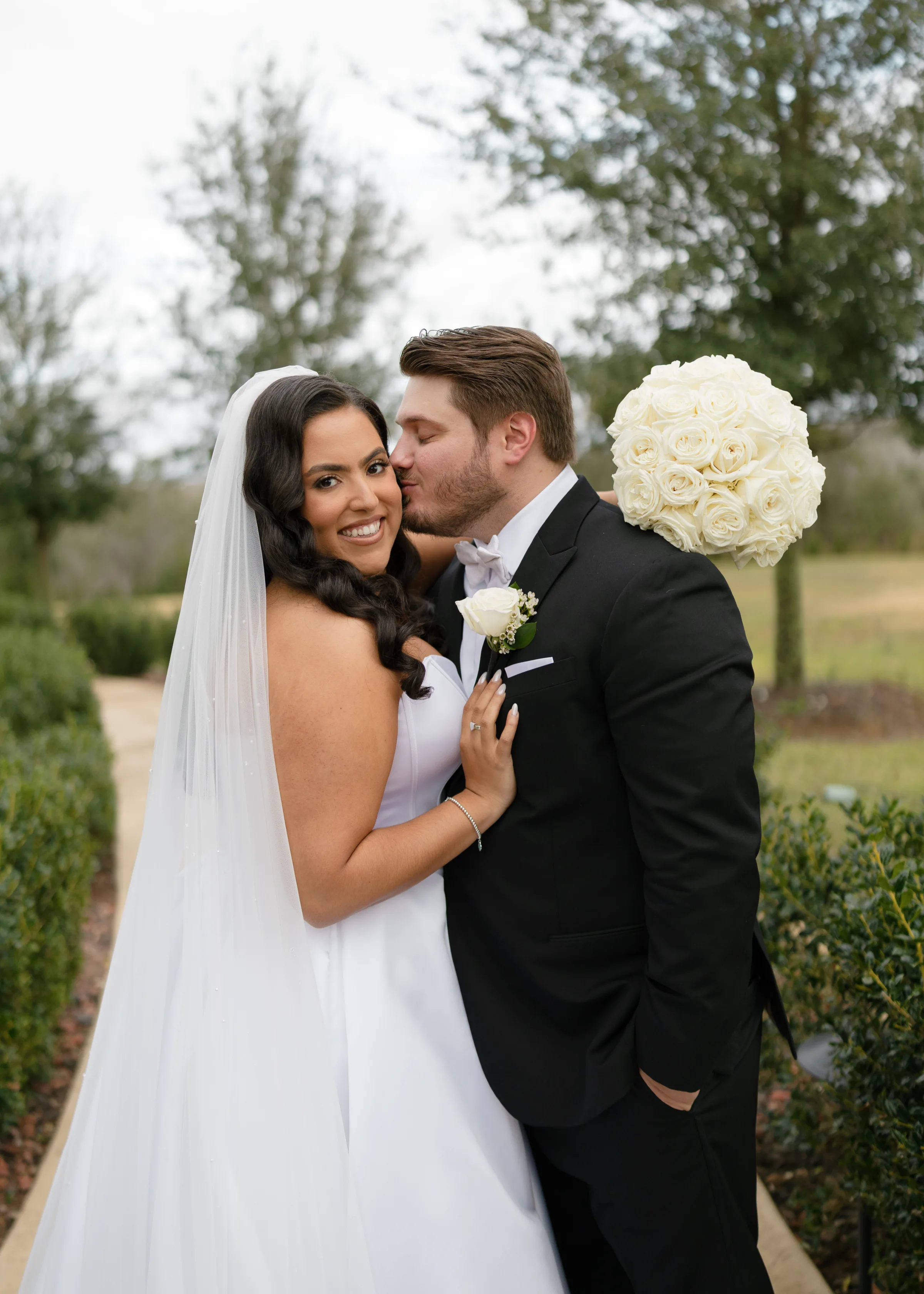 Bride in strapless white gown and veil smiles as groom in black tuxedo kisses her cheek while she holds a lush white rose bouquet in an outdoor garden setting in Central Florida