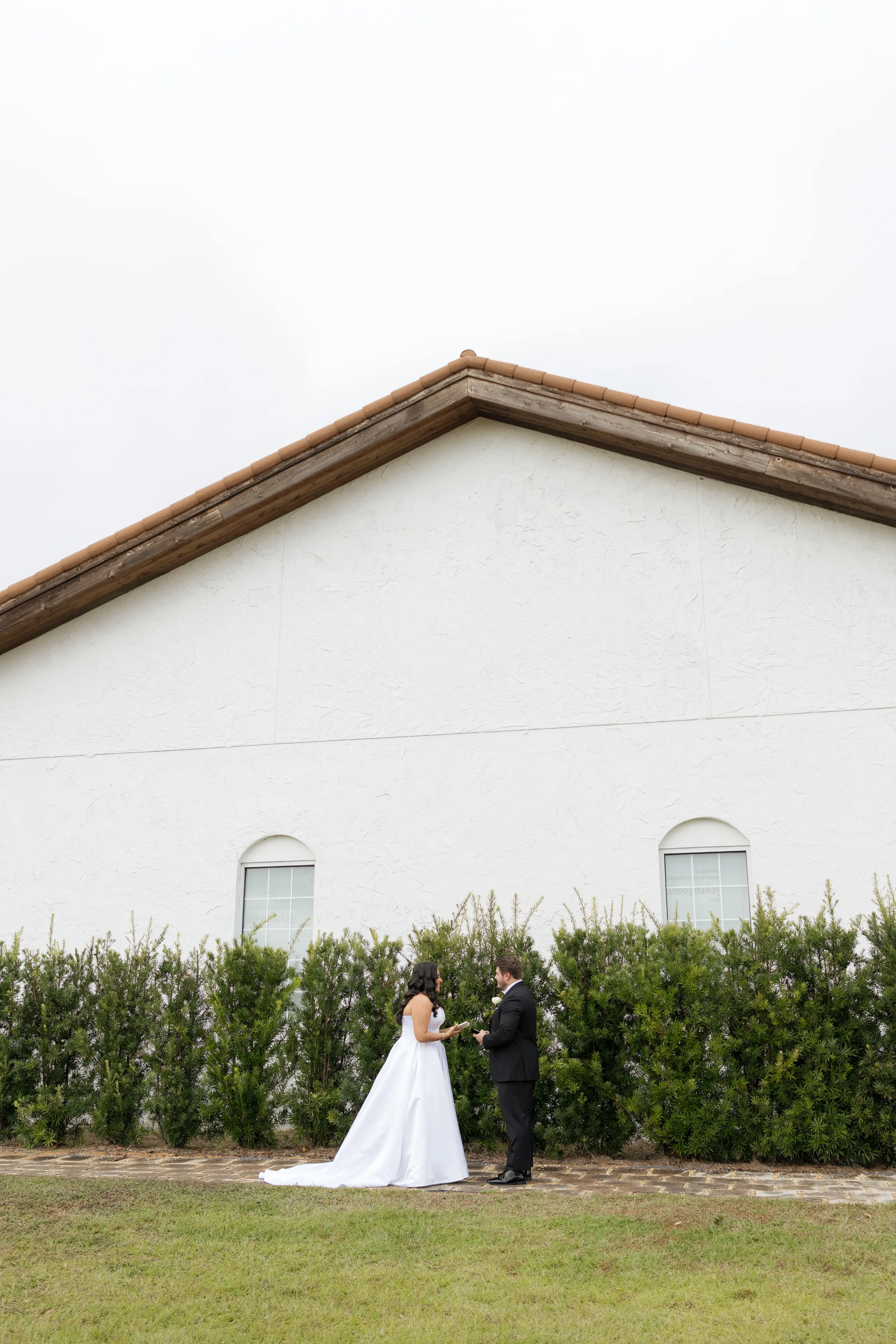 Bride in white gown and groom in black tuxedo share a private vow reading moment outside a white barn wedding venue in Central Florida