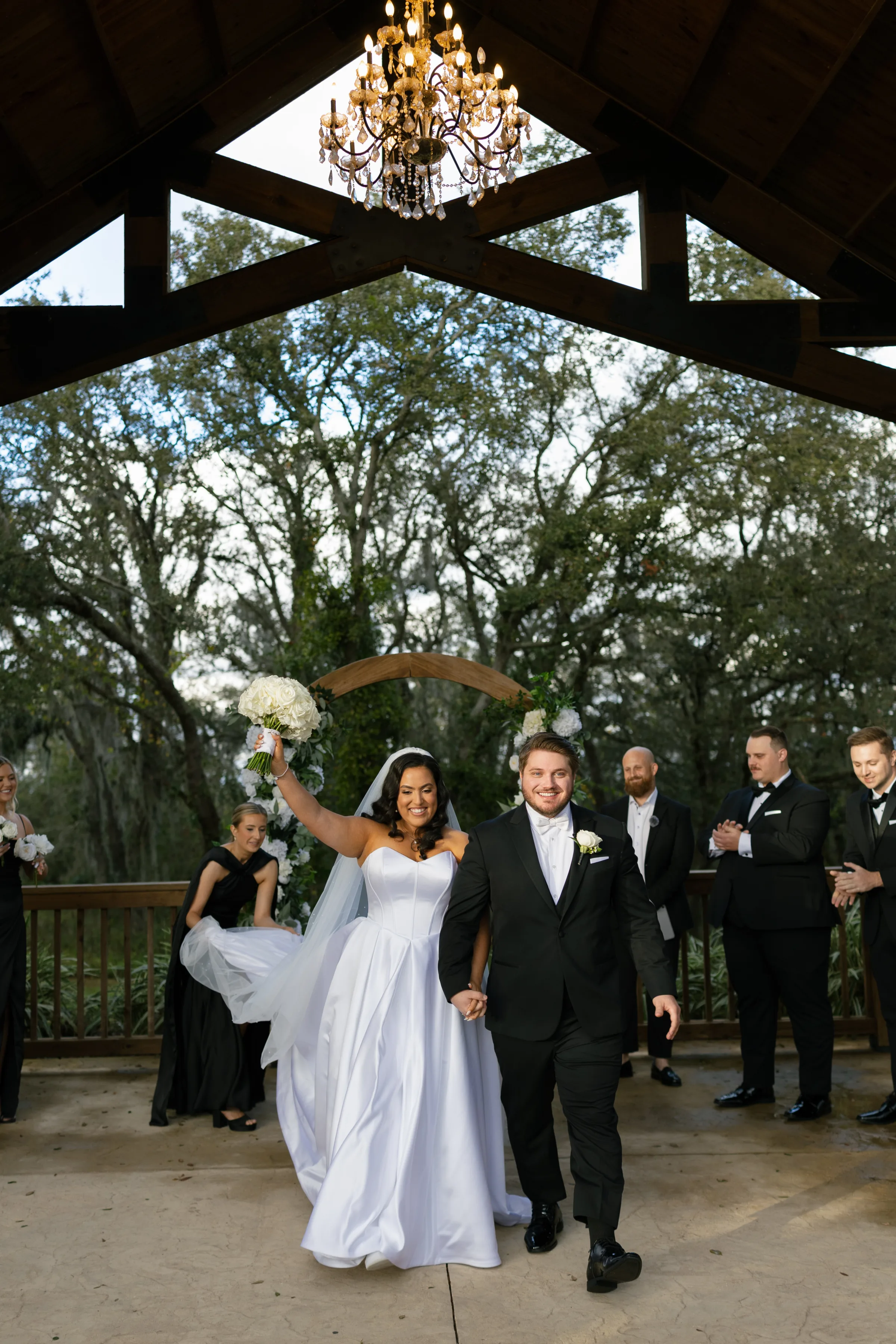 Bride raising bouquet of white roses while walking the recessional with her groom under a chandelier-lit pavilion surrounded by wedding party at a Central Florida outdoor wedding venue