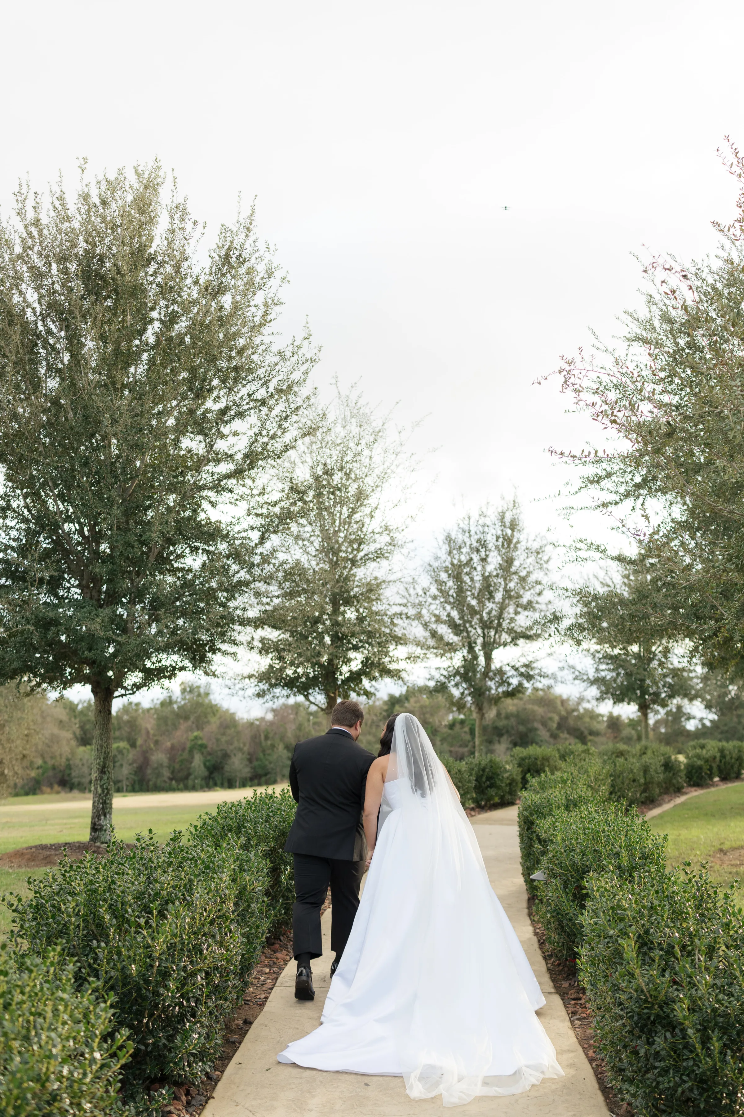 Bride in a white gown and veil walks hand in hand with groom in black tuxedo along a manicured garden pathway at an Orlando Central Florida wedding venue