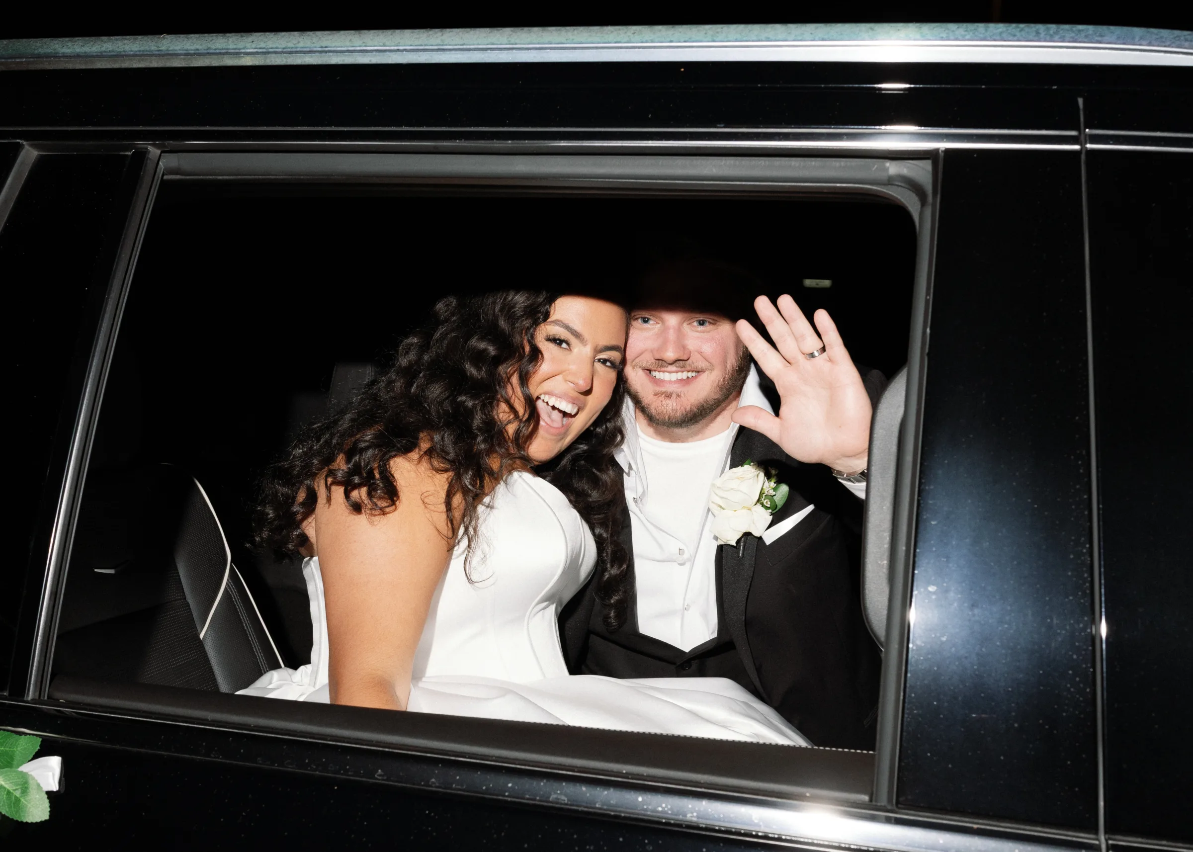 Smiling bride and groom waving from a black luxury car after their Central Florida wedding ceremony, groom wearing a boutonniere and wedding band