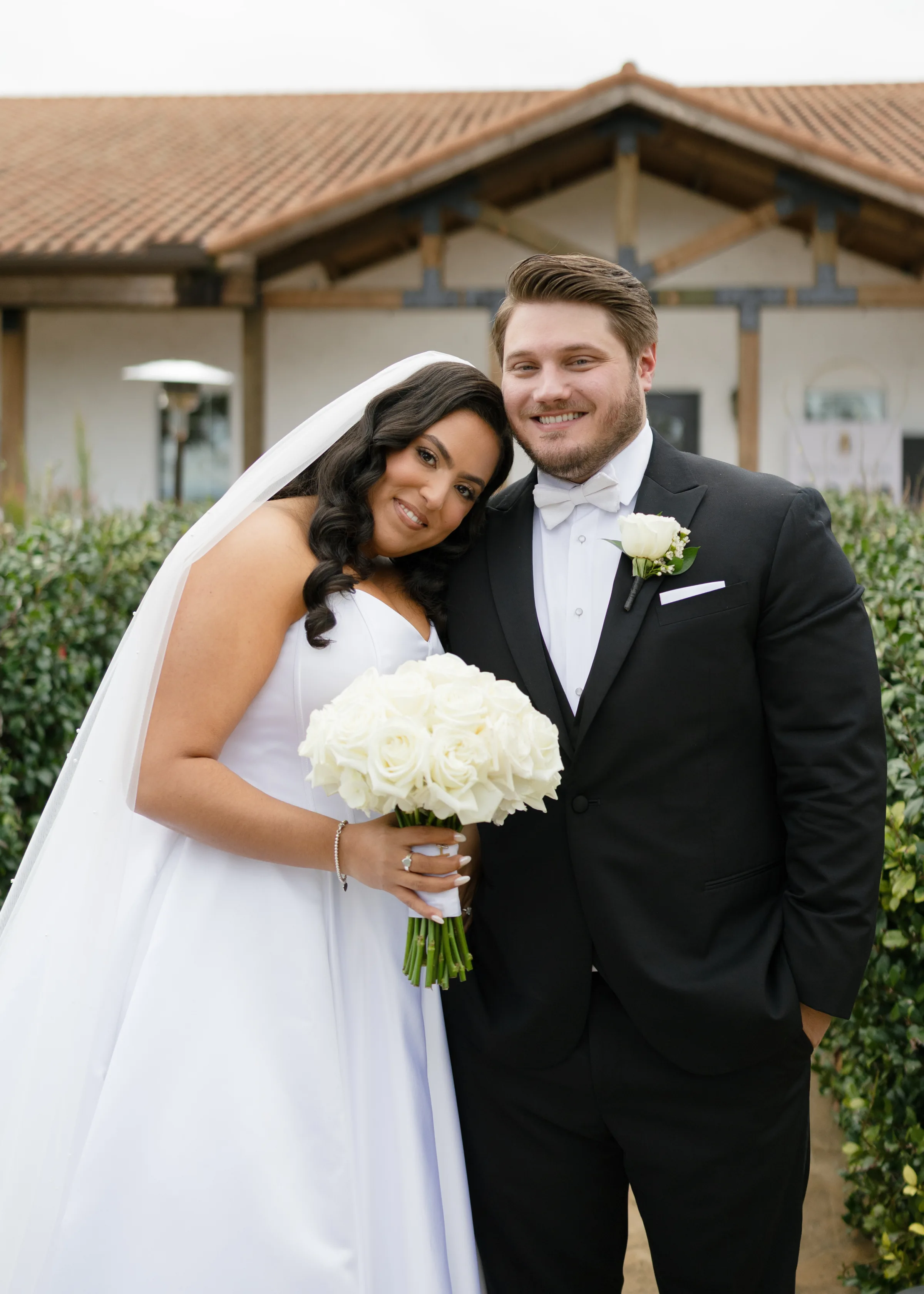Smiling bride in white gown and veil holding a white rose bouquet leans against groom in black tuxedo with white bow tie and boutonniere at an Orlando Central Florida wedding venue