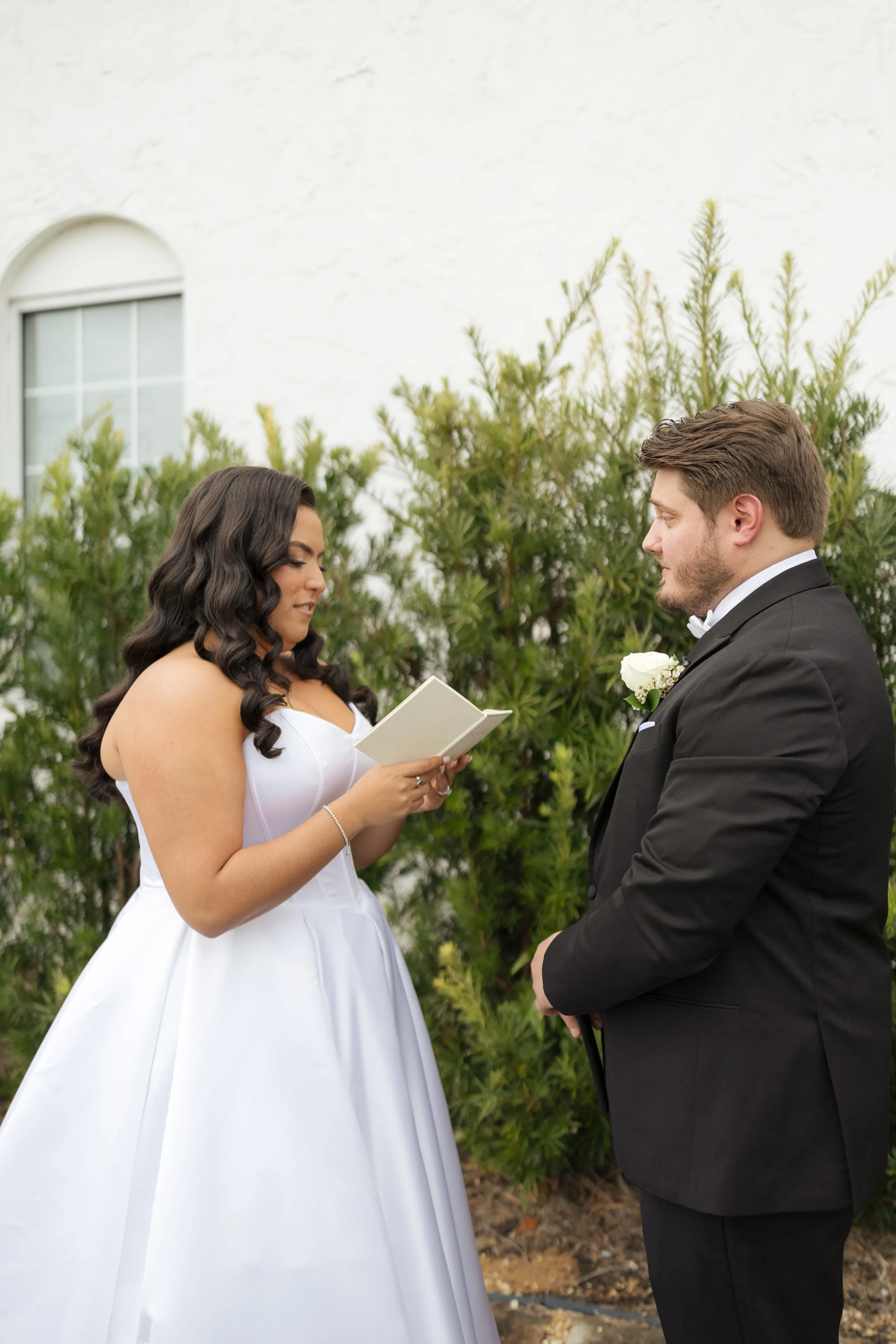 Bride in a strapless white gown reads personal vows from a small book to her groom in a black tuxedo during an outdoor wedding ceremony in Central Florida