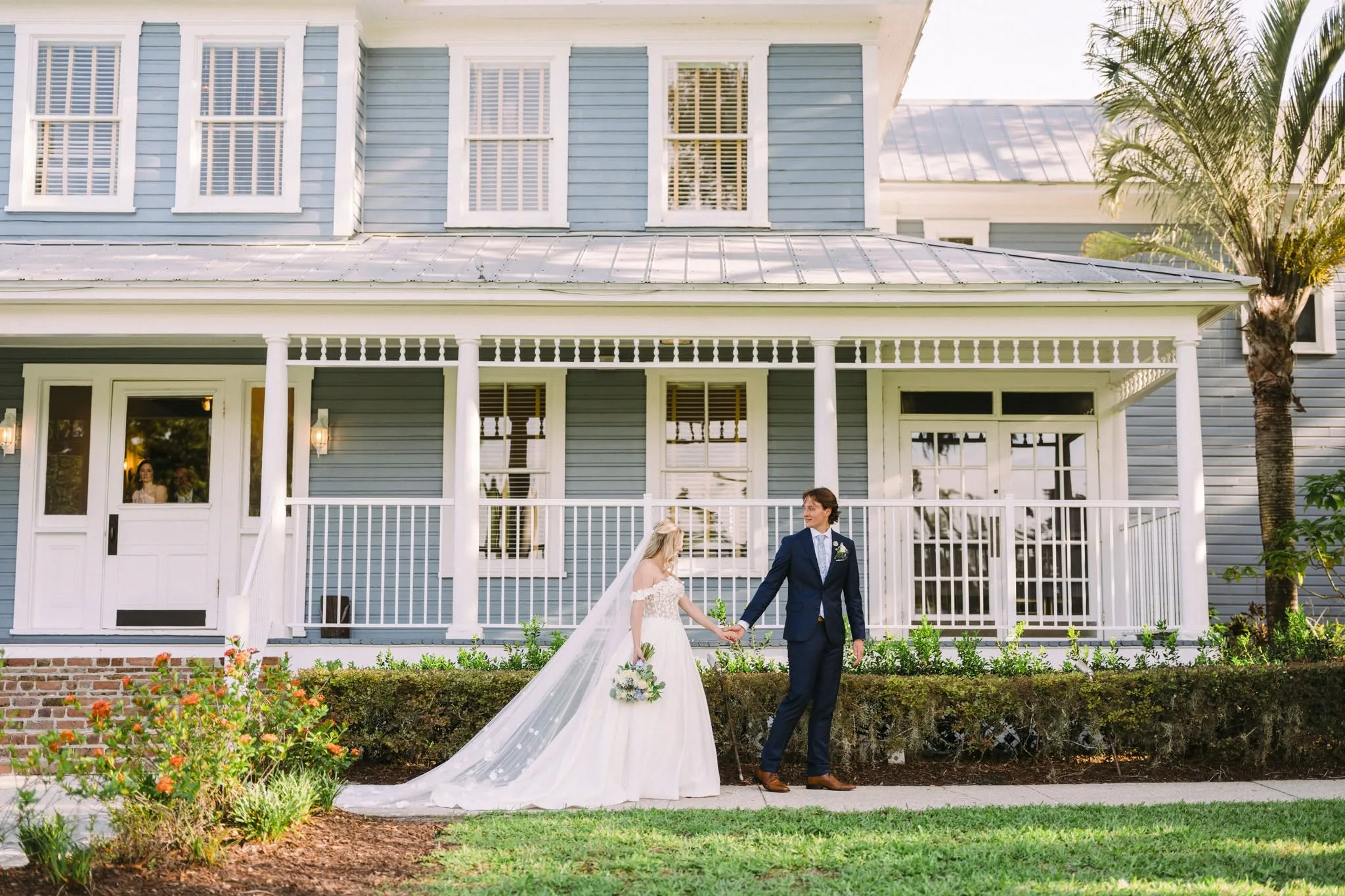 Bride in off-shoulder gown and groom in navy suit hold hands on the lawn in front of a blue colonial wedding venue in Central Florida
