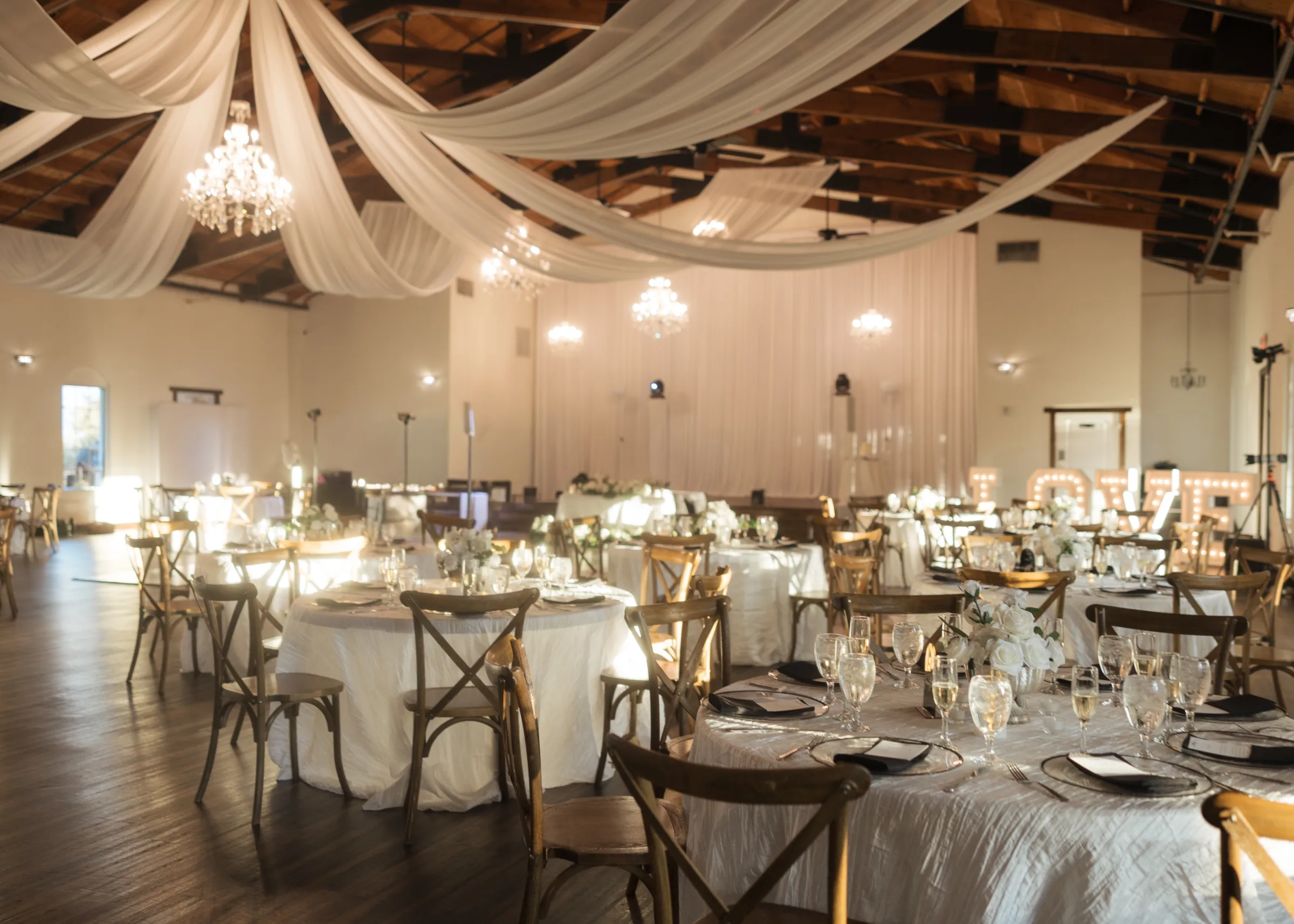 Elegantly decorated wedding reception hall in Orlando Central Florida featuring white ceiling draping, crystal chandeliers, rustic cross-back chairs, and white floral centerpieces on round tables with glowing marquee letters in the background