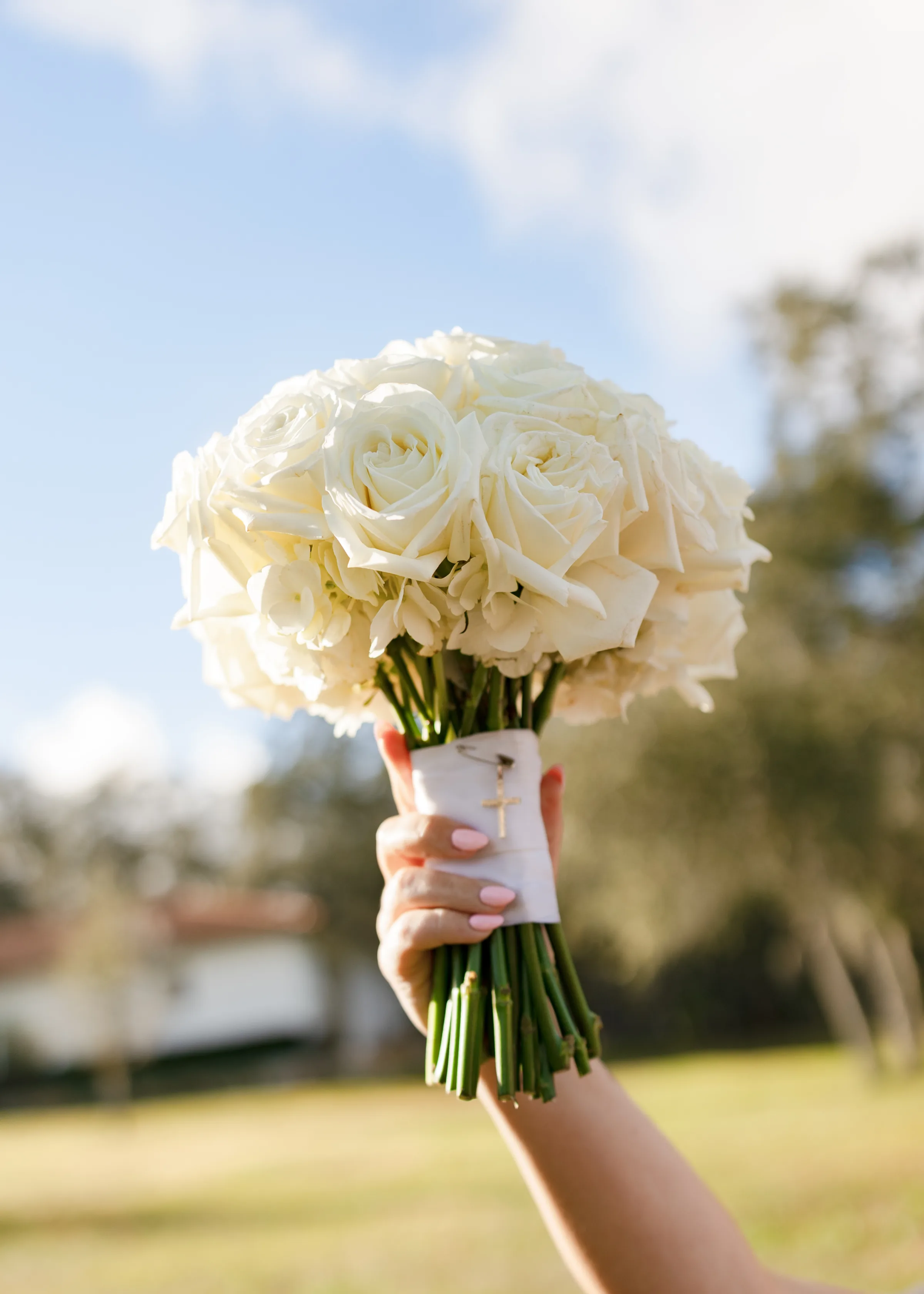 A hand holds a classic all-white rose bridal bouquet with a gold cross charm wrapped at the stems, photographed outdoors at a Central Florida wedding venue