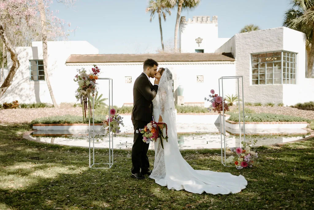 garden venue AH Museum Maitland Joanna Moore Photo 202577 - Wedding Venues Collections Bride and groom sharing their first kiss during an outdoor garden wedding ceremony at a white Spanish-style Art History Museum of Maitland in Central Florida, surrounded by colorful floral arch arrangements on modern geometric metal stands with a reflective pond in the background