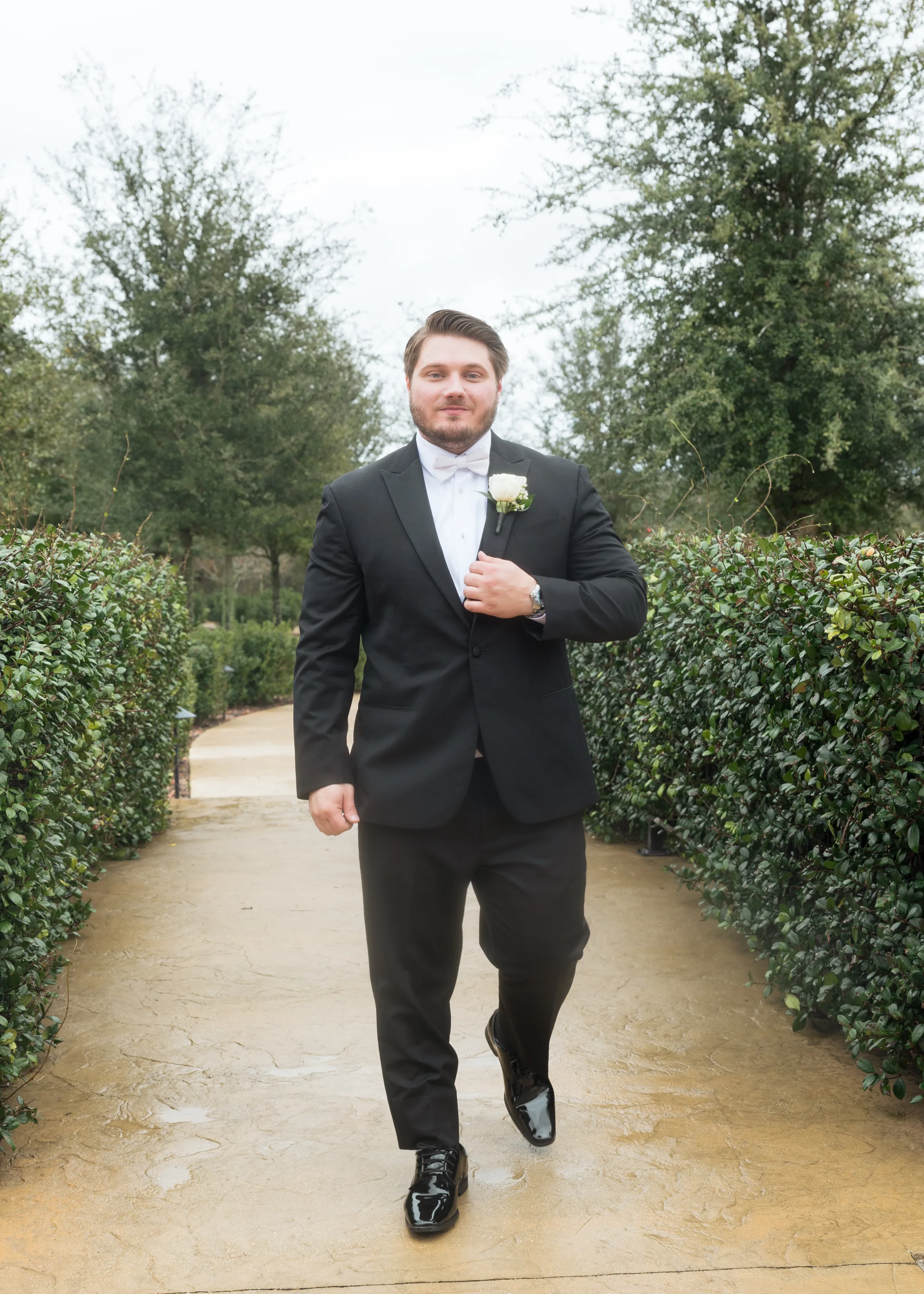 Groom in a black tuxedo with white bow tie and rose boutonniere walking confidently down a hedged garden pathway at a Central Florida wedding venue