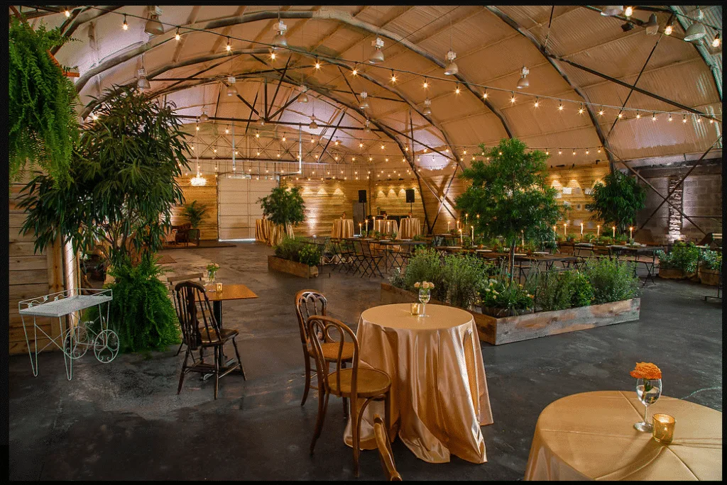 industrial central florida wedding venue 1010 west - Wedding Venues Collections Industrial wedding venue interior with arched ceiling, string lights, lush greenery garden beds, gold satin cocktail tables, and warm candlelight ambiance at 1010 West in Central Florida
