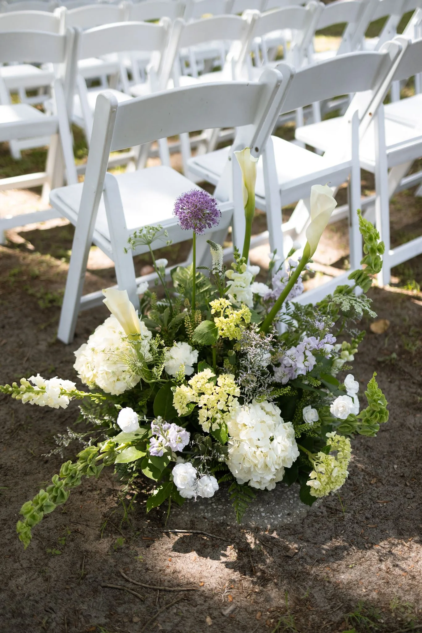 lilac ivory wedding aisle florals la hacienda snow hill - An Elegant Lilac and Ivory Wedding at La Hacienda on Snow Hill Lilac and ivory wedding aisle floral arrangement featuring white hydrangeas, calla lilies, and purple allium beside white folding chairs at La Hacienda on Snow Hill in Central Florida