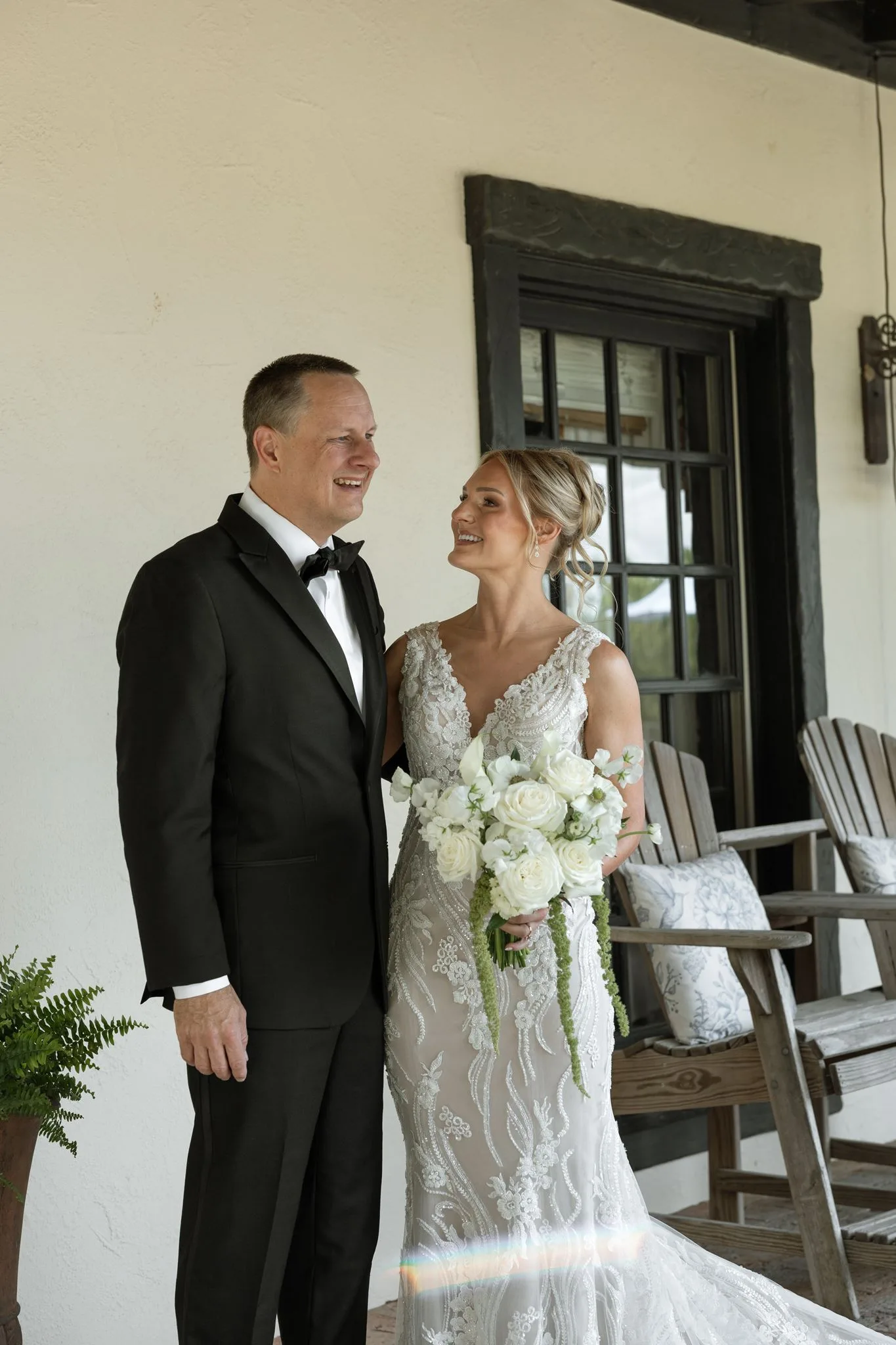 lilac ivory wedding bride groom la hacienda snow hill 1 - An Elegant Lilac and Ivory Wedding at La Hacienda on Snow Hill Bride in lace fitted gown holding white rose bouquet smiles at groom in black tuxedo on the porch of La Hacienda on Snow Hill in Orlando Florida for their lilac and ivory spring wedding