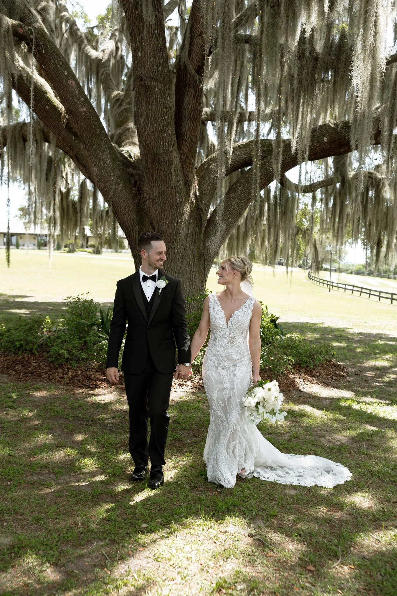 lilac ivory wedding bride groom la hacienda snow hill - An Elegant Lilac and Ivory Wedding at La Hacienda on Snow Hill Bride and groom holding hands and smiling beneath a sprawling Spanish moss oak tree at La Hacienda on Snow Hill in Central Florida, bride wearing a fitted lace gown and holding a white bouquet, groom in a classic black tuxedo.