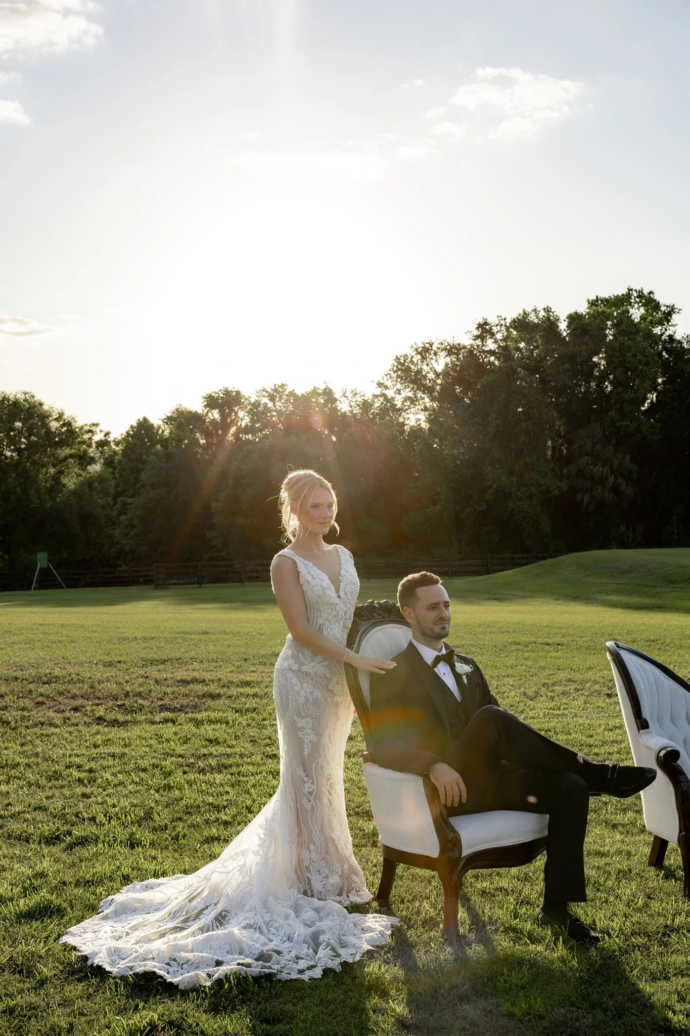 lilac ivory wedding bride groom portrait la hacienda - An Elegant Lilac and Ivory Wedding at La Hacienda on Snow Hill Bride in lace gown standing beside seated groom in tuxedo during golden hour sunset portrait at La Hacienda on Snow Hill in Central Florida for a lilac and ivory wedding celebration