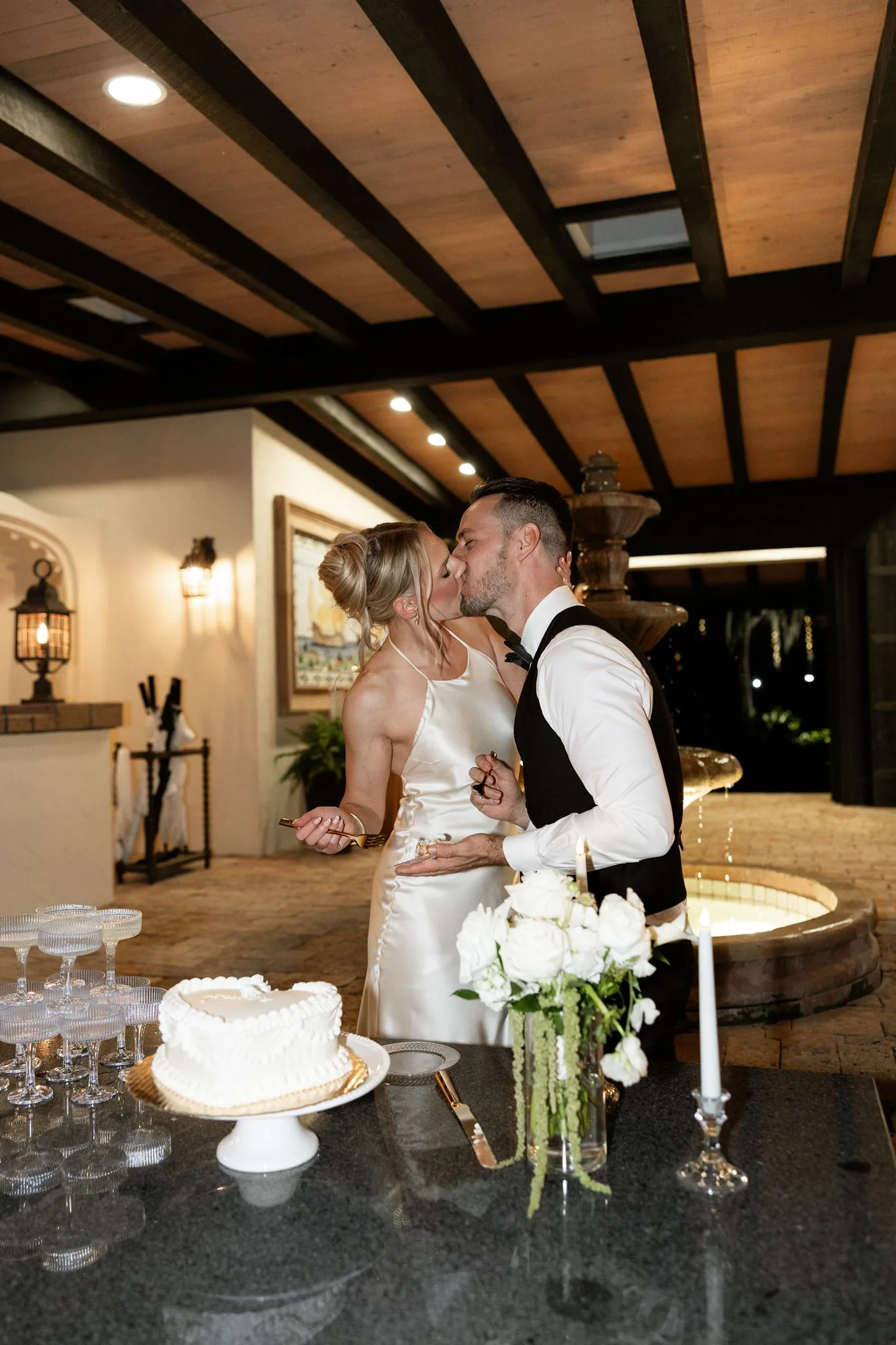 lilac ivory wedding cake cutting la hacienda snow hill - An Elegant Lilac and Ivory Wedding at La Hacienda on Snow Hill Bride and groom sharing a kiss while cutting their white heart-shaped wedding cake at La Hacienda on Snow Hill in Orlando, Florida, surrounded by elegant champagne coupes and white floral arrangements in a romantic Mediterranean-inspired venue setting