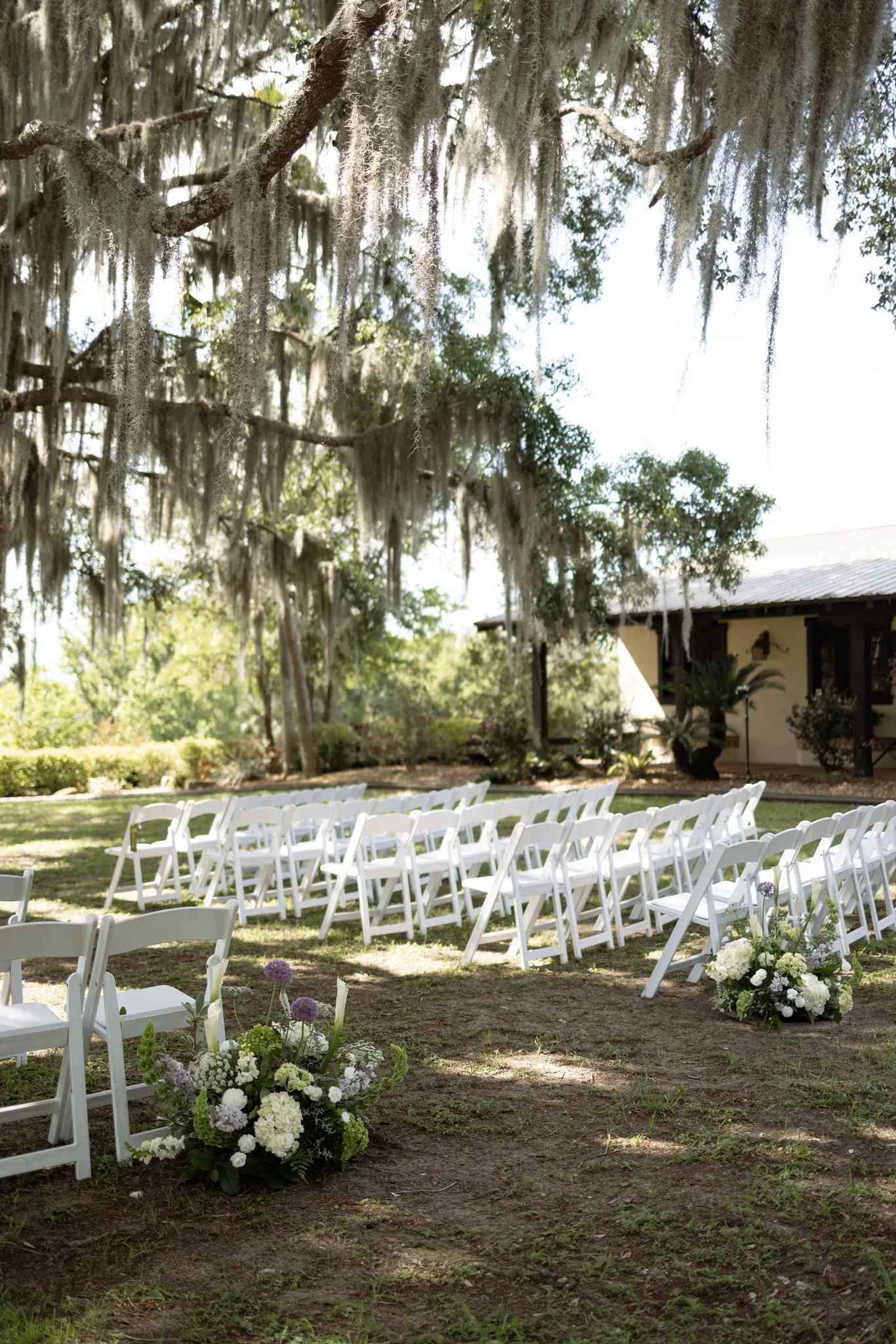 lilac ivory wedding ceremony la hacienda snow hill - An Elegant Lilac and Ivory Wedding at La Hacienda on Snow Hill Outdoor lilac and ivory wedding ceremony setup with white folding chairs, lush floral arrangements, and Spanish moss-draped oak trees at La Hacienda on Snow Hill in Central Florida