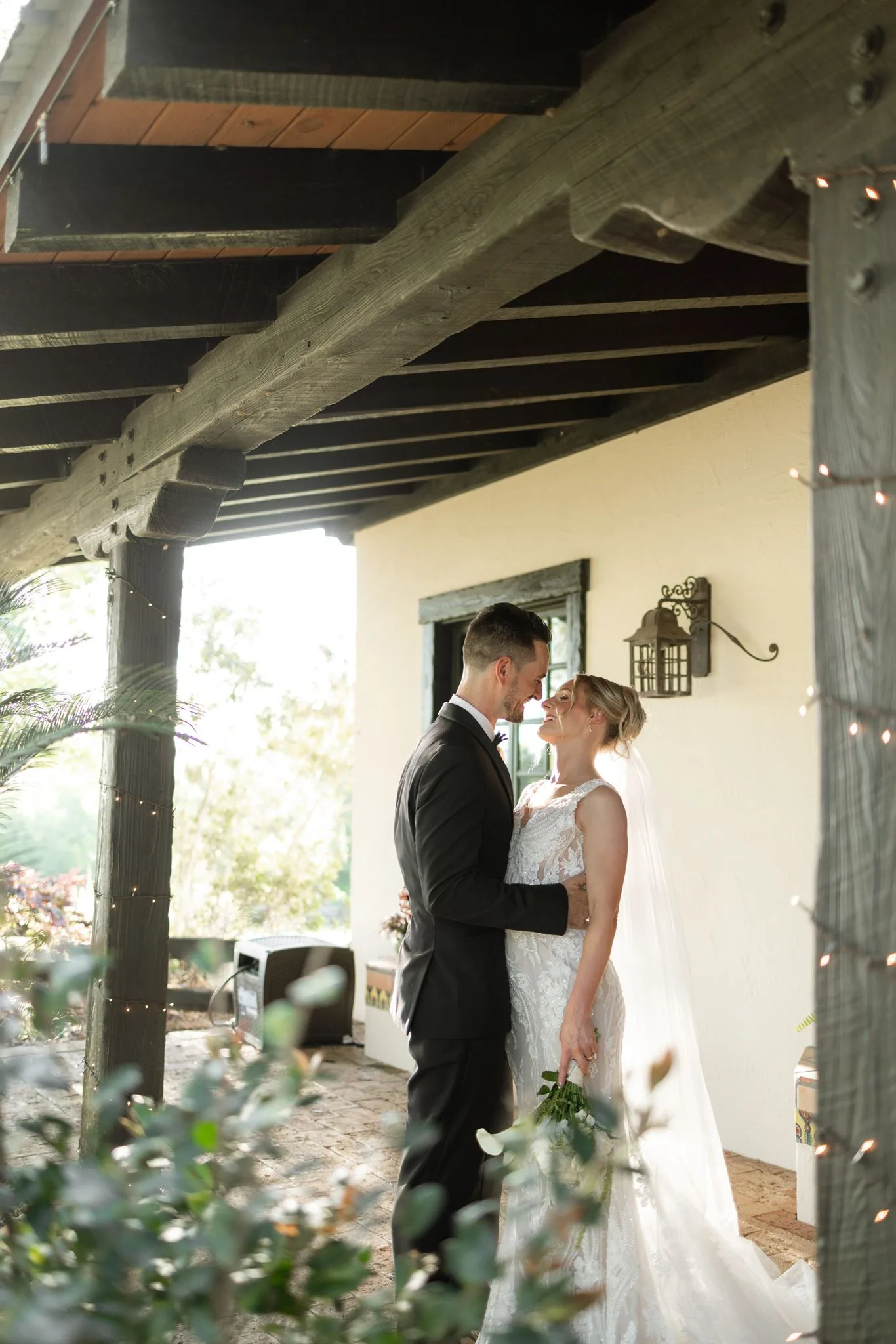 lilac ivory wedding couple la hacienda snow hill 1 - An Elegant Lilac and Ivory Wedding at La Hacienda on Snow Hill Bride and groom share an intimate moment under rustic wood beam pergola at La Hacienda on Snow Hill wedding venue in Central Florida, bride in lace gown holding greenery bouquet during lilac and ivory spring wedding celebration