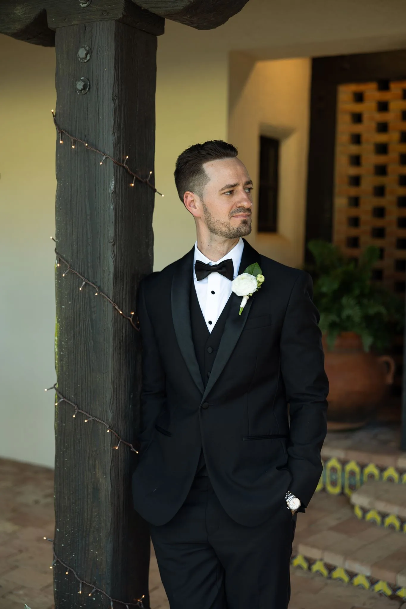 lilac ivory wedding groom tuxedo la hacienda snow hill - An Elegant Lilac and Ivory Wedding at La Hacienda on Snow Hill Groom in black tuxedo with white boutonniere leaning against a rustic wooden pillar at La Hacienda on Snow Hill in Orlando Florida for a lilac and ivory wedding celebration