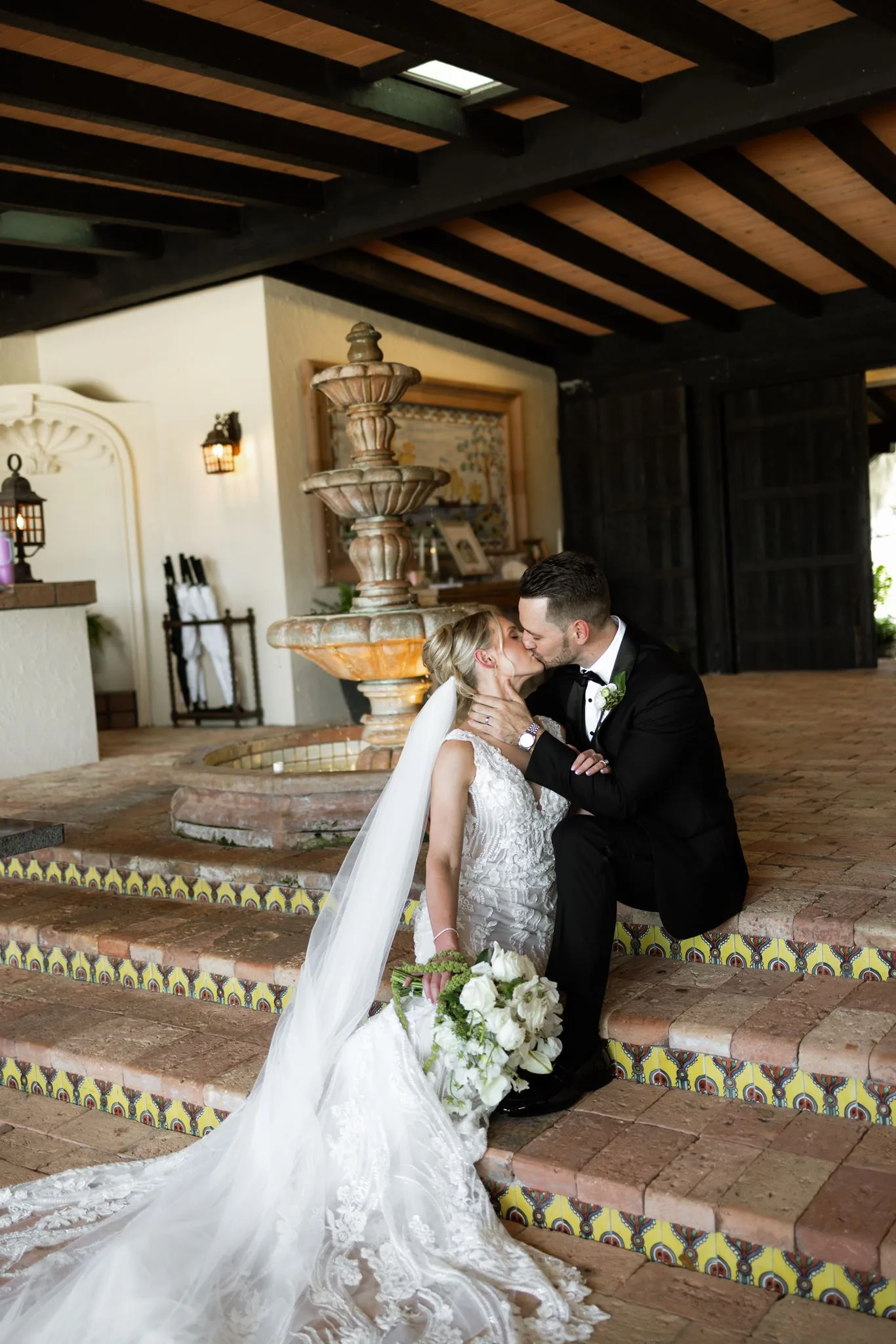 lilac ivory wedding kiss la hacienda snow hill - An Elegant Lilac and Ivory Wedding at La Hacienda on Snow Hill Bride and groom share a romantic kiss beside a Spanish-style fountain at La Hacienda on Snow Hill in Orlando, Florida, with the bride holding a white floral bouquet in her lace wedding gown