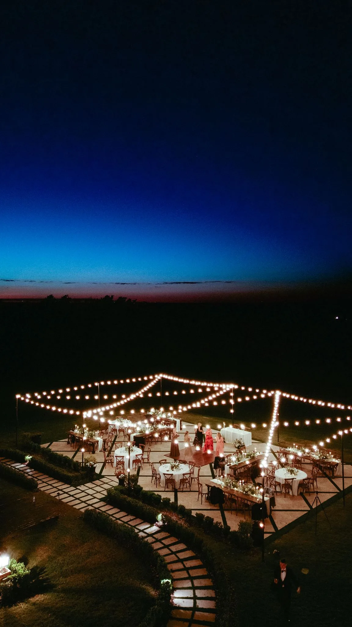 lilac ivory wedding reception la hacienda snow hill - An Elegant Lilac and Ivory Wedding at La Hacienda on Snow Hill Aerial twilight view of an elegant outdoor wedding reception at La Hacienda on Snow Hill in Central Florida, featuring string lights canopy over round and farm tables with lilac and ivory floral centerpieces and a dance floor glowing against a deep blue sunset sky