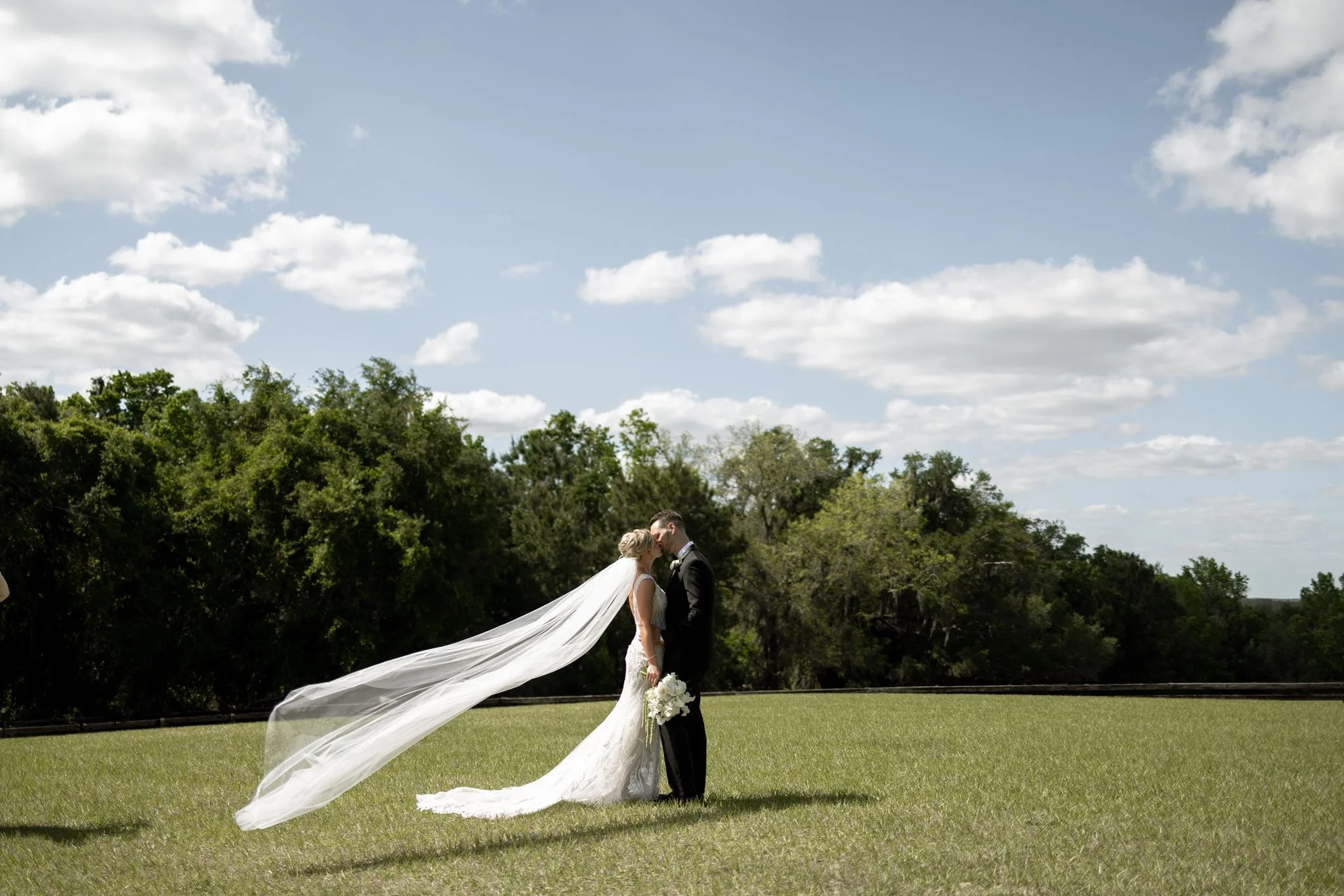 lilac ivory wedding venue la hacienda snow hill - An Elegant Lilac and Ivory Wedding at La Hacienda on Snow Hill Bride and groom sharing a first kiss on the open lawn at La Hacienda on Snow Hill in Central Florida, with the bride's cathedral veil flowing dramatically in the wind against a blue sky backdrop.