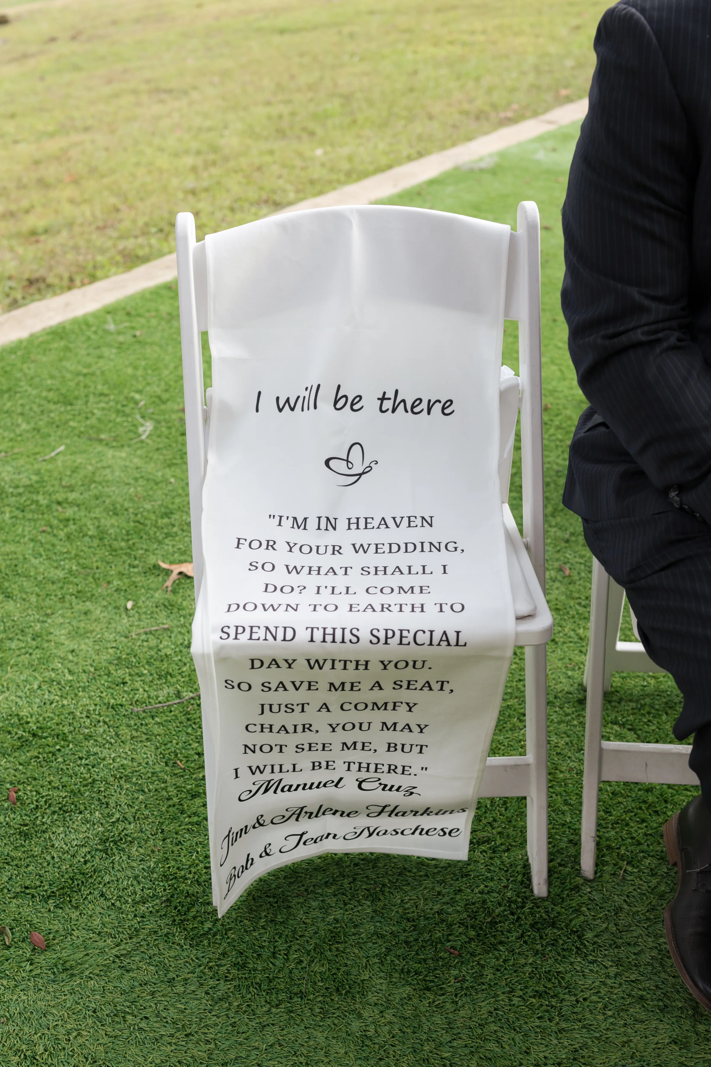 A white folding memorial chair with a printed sash reading 'I Will Be There' honoring deceased loved ones Manuel Cruz, Jim and Arlene Harkins, and Bob and Jean Noschese at an outdoor wedding ceremony in Central Florida