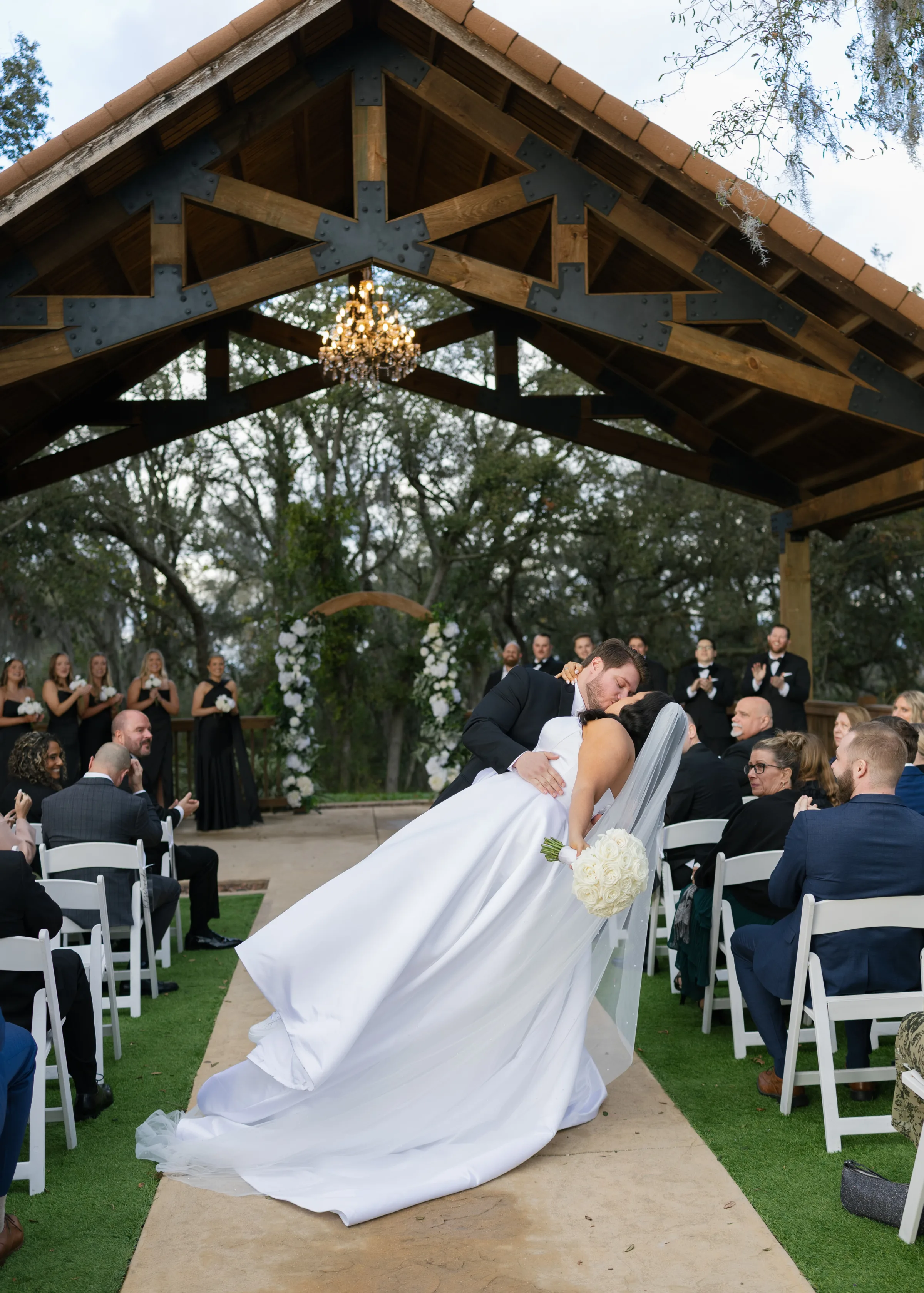Bride and groom share a romantic dip kiss in the aisle during their outdoor wedding ceremony under a rustic wooden pavilion with chandelier in Central Florida