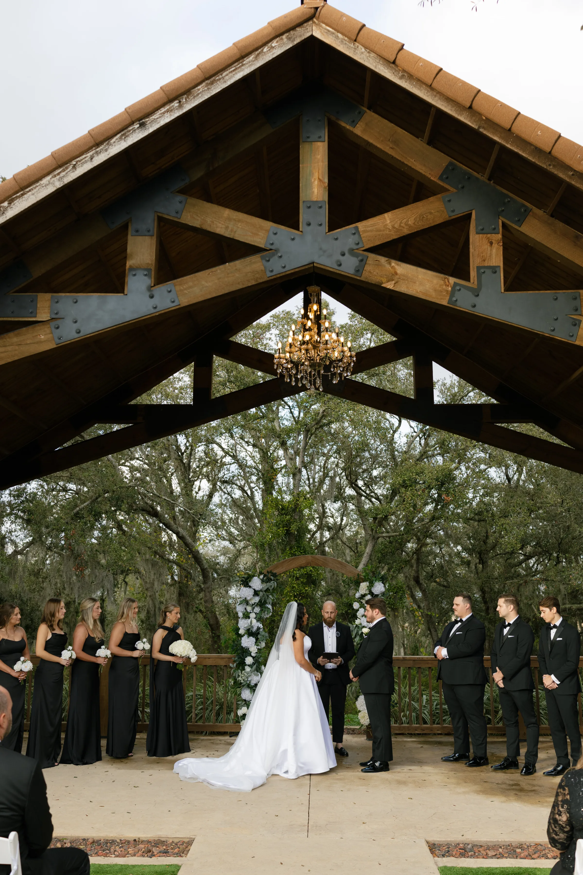 Bride and groom exchange vows under a rustic wood pavilion chandelier with bridesmaids in black gowns and groomsmen in tuxedos surrounded by white floral arrangements and oak trees at a Central Florida wedding venue