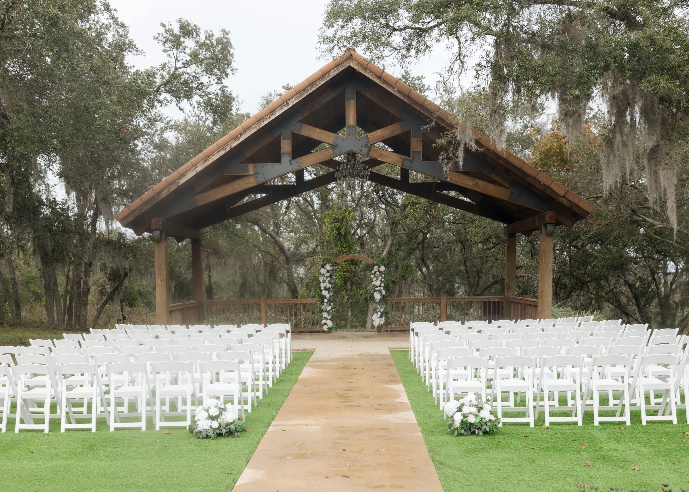 Rustic wooden pavilion wedding ceremony setup with white folding chairs lining a grass aisle, floral arch, and Spanish moss-draped oak trees at an outdoor Central Florida wedding venue