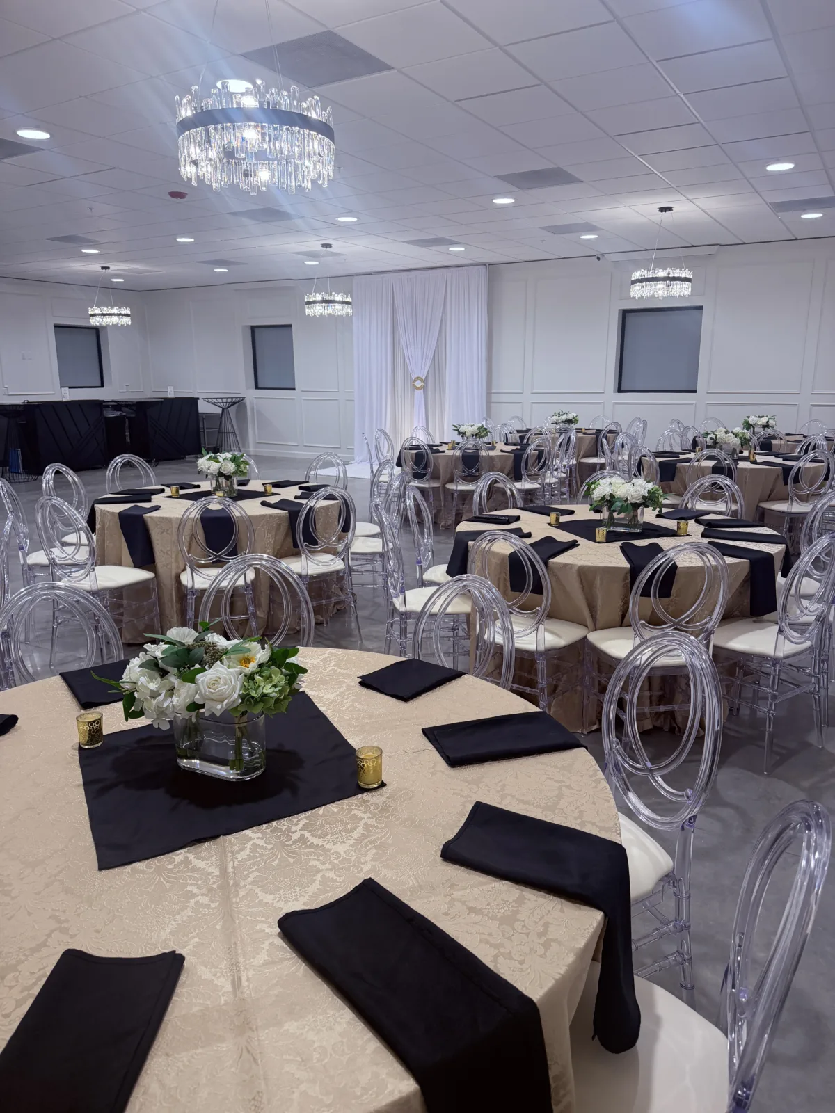Elegant ballroom reception setup at Platinum Celebrations Event Center in Apopka Florida featuring clear acrylic oval-back chairs, champagne damask tablecloths, black napkins, white floral centerpieces, and crystal chandeliers in a luxe timeless wedding venue style