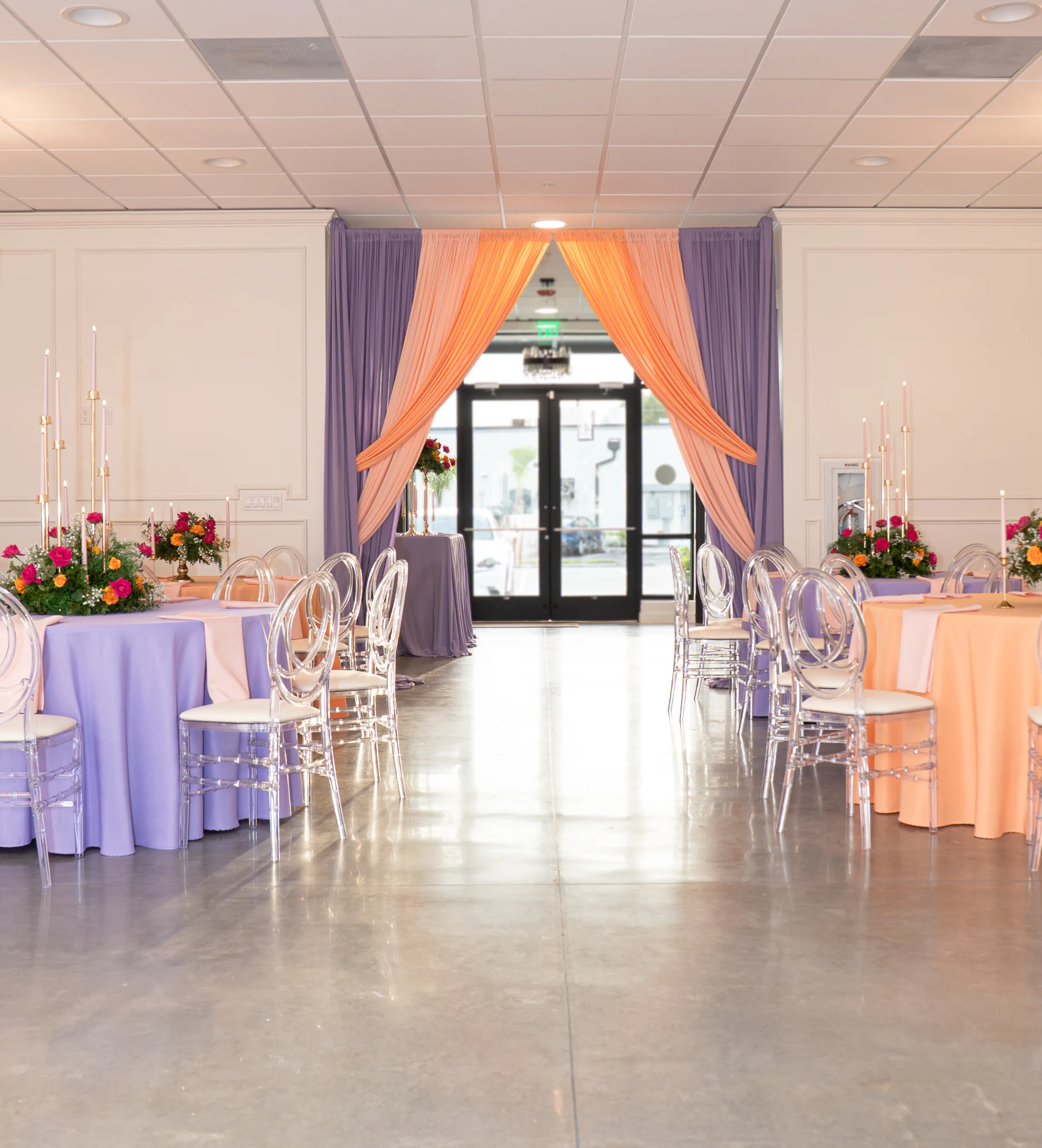 Elegant wedding reception setup at Platinum Celebrations Event Center in Apopka Florida featuring lavender and peach tablecloths on round tables with clear acrylic ghost chairs tall gold candelabras colorful floral centerpieces and a draped peach and purple fabric backdrop framing the entrance doors