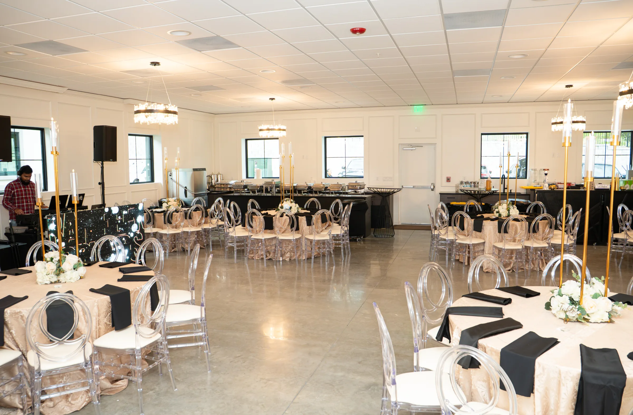 Elegant ballroom reception setup at Platinum Celebrations Event Center in Apopka Florida featuring round tables with champagne linen tablecloths, black napkins, acrylic ghost chairs, tall gold candelabra centerpieces with white floral arrangements, crystal chandeliers, and a buffet station along the back wall