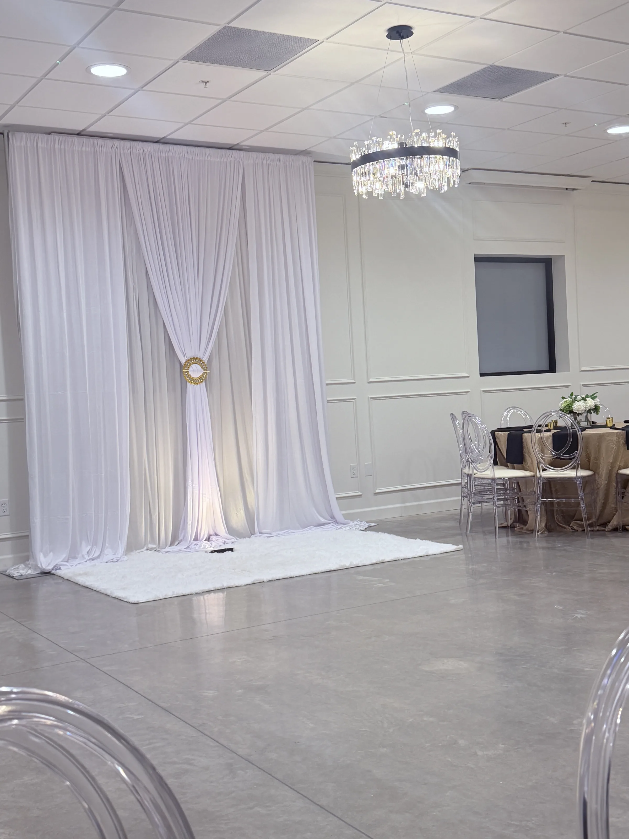 White draped fabric backdrop with gold sunburst tie-back accent on a white rug inside the elegant ballroom at Platinum Celebrations Event Center in Apopka Florida, styled with ghost chairs and gold and black table linens for a luxe wedding reception