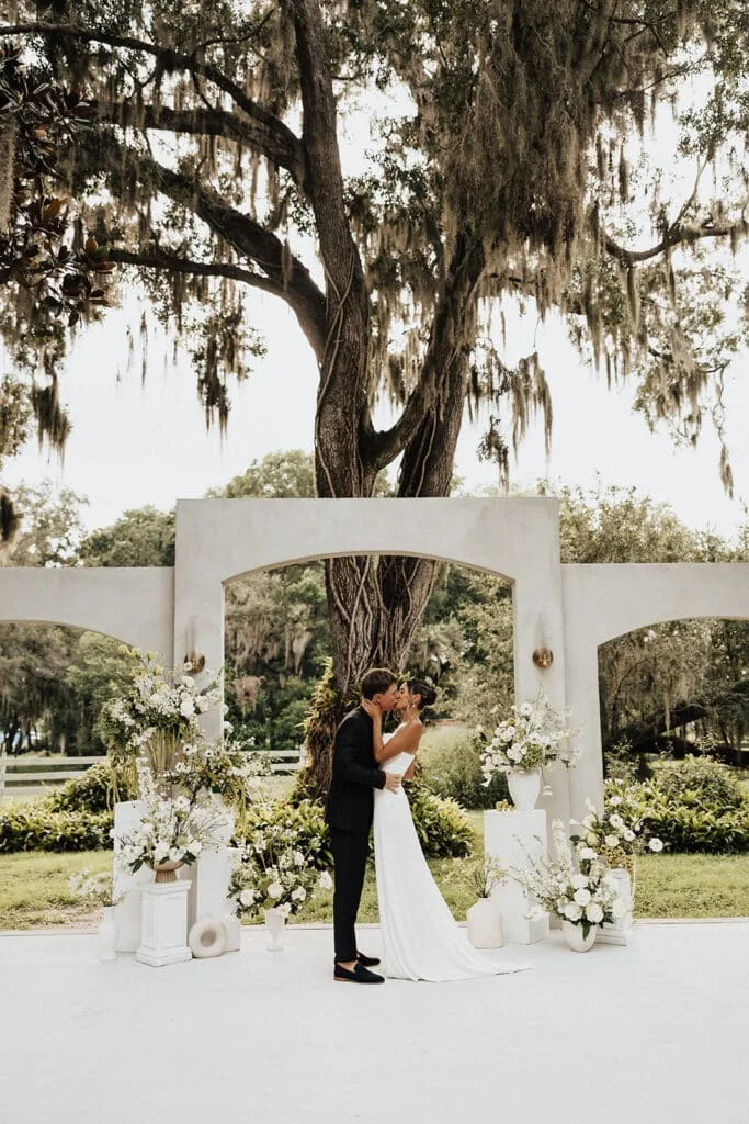 romantic central florida wedding venue uliana estate - Wedding Venues Collections bride and groom sharing first kiss under a grand live oak tree draped in Spanish moss at an elegant outdoor wedding ceremony at Uliana Estate wedding venue in Central Florida, framed by a white arch altar surrounded by white floral arrangements